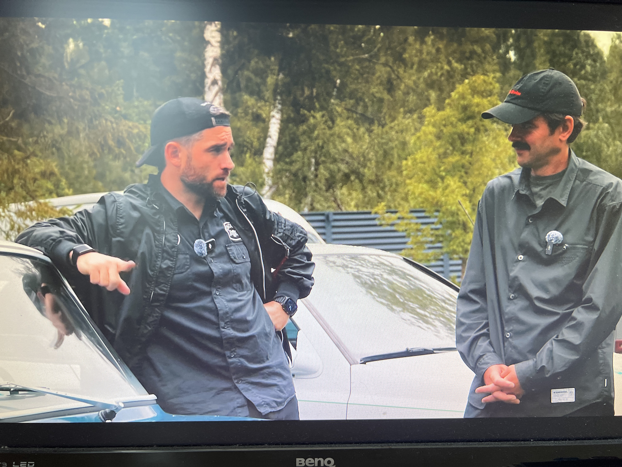 Two men are standing outdoors near cars. The man on the left is wearing a black baseball cap, a black bomber jacket over a black button-up shirt, and dark pants. He has a beard and is leaning on a car, pointing with his left hand. He is wearing a watch on his left wrist. The man on the right is wearing a black baseball cap with red lettering, a dark button-up shirt, and dark pants. He has a mustache. He is standing with his hands clasped in front of him. Both men have pins on their shirts. In the background, there are trees and a metal fence. The scene appears to be in an outdoor area, possibly a yard or parking lot. There is no information in the image to suggest a specific location in the city of Kanash, Russia.