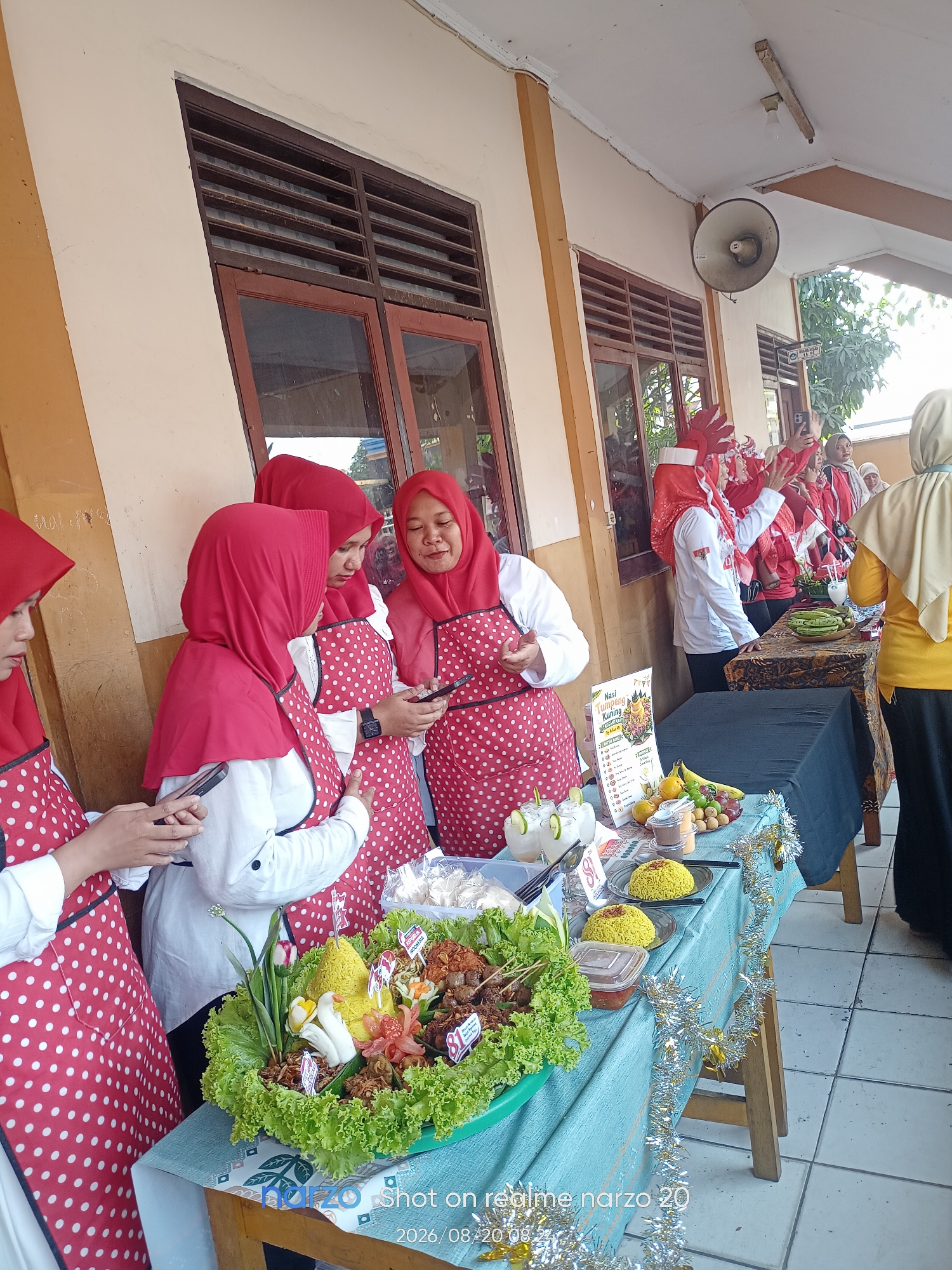 This image captures an outdoor food stall event, likely at a school or community gathering, in Karawang, Indonesia. The scene is set against a building with a light-colored facade and wooden-framed windows with horizontal louvers. Four women, dressed in red hijabs and red aprons with white polka dots over white shirts, are the central focus. Two of them are actively engaged with their mobile phones, while another appears to be looking at her device. A fourth woman, standing further back, is also looking down.
On the tables, a vibrant display of food is presented. A large green platter is filled with a colorful arrangement of food items, including yellow rice shaped into a cone, skewers of meat, shredded chicken, and garnished with lettuce, tomatoes, and small Indonesian flags. Adjacent to this, two plates of yellow rice are set out, along with a small container of chili sauce. A sign with Indonesian text, which appears to be a menu, is also visible. Further along the table, there are fruits such as bananas, grapes, and limes, as well as glasses of what seems to be a refreshing beverage. Decorative silver tinsel is draped across the table.
In the background, several other individuals are present, some also wearing red head coverings. One person in the background appears to be holding a small Indonesian flag and waving. A speaker is mounted on the wall above the stall. The floor is tiled with a light-colored, square pattern. The overall atmosphere suggests a lively, communal event, possibly a culinary exhibition or a fundraising activity.