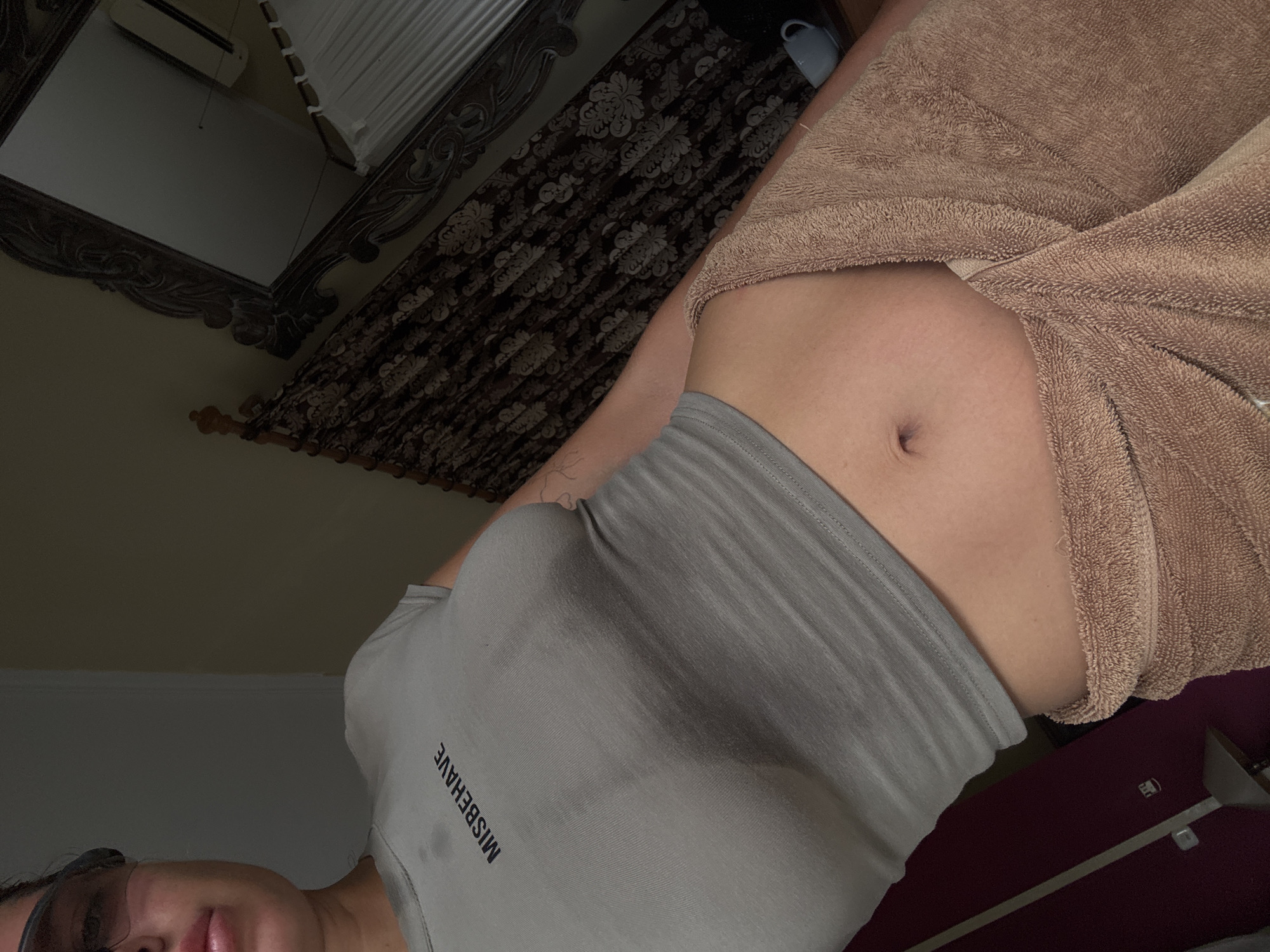 The image presents a partial view of a person indoors. They are wearing a gray tank top with the word "MISBEHAVE" printed vertically on the left side and a tan towel wrapped around their waist. Their midriff is exposed. A portion of their face, including glasses and lips, is visible in the lower left. The background includes a dark, ornate headboard, part of a mirror reflecting an air conditioning unit and a window with closed blinds. The environment appears to be a bedroom, although its exact location within Unknown, Indonesia, cannot be determined from the provided image.