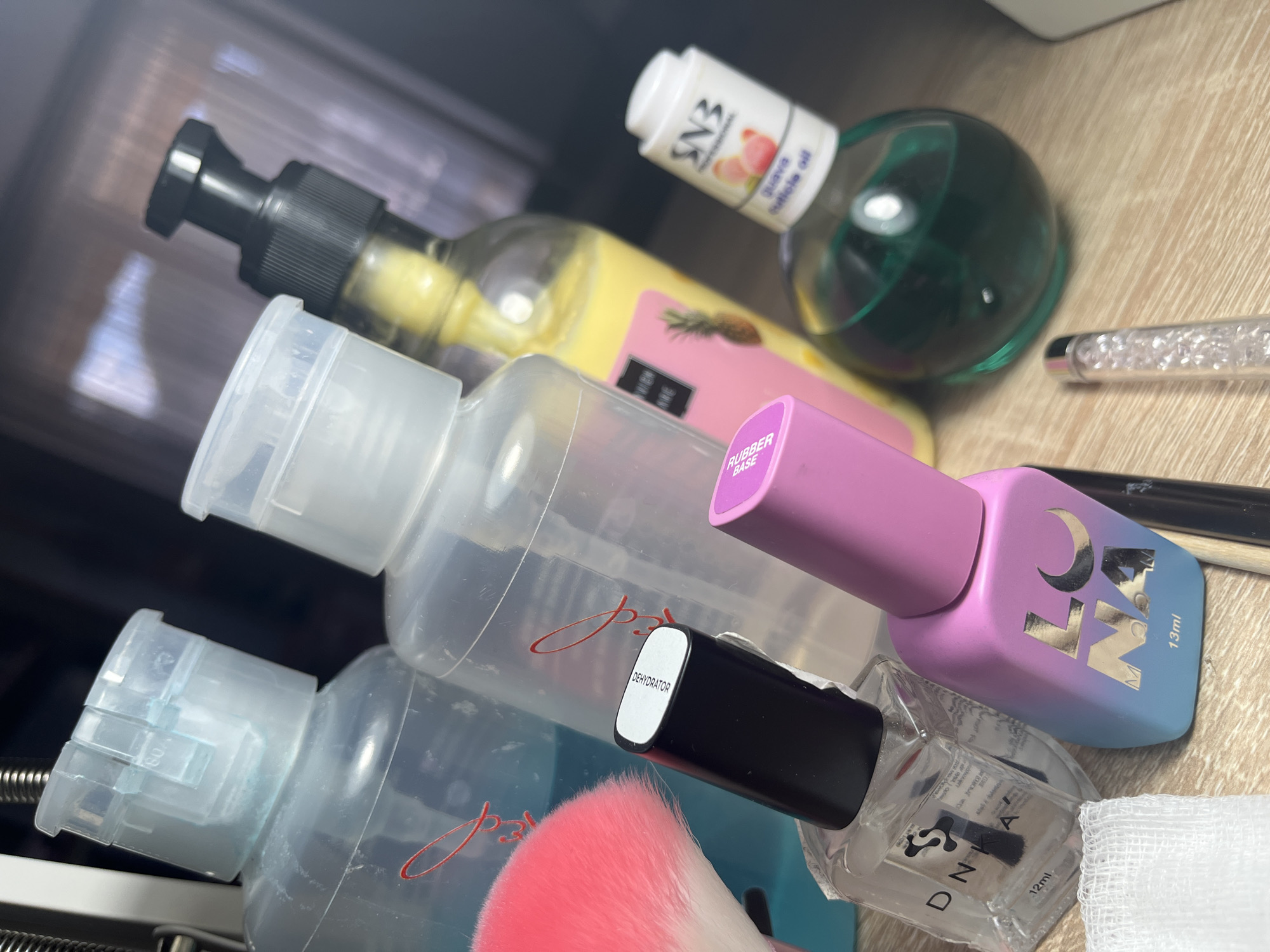 The image shows a cluttered collection of nail care products arranged on a light wooden surface. Prominently featured are several bottles: one clear plastic bottle with a white cap and a barely visible red script, another similar bottle containing a blue liquid, and a third clear plastic bottle with a clear cap. Next to these is a small bottle labeled "DEHYDRATOR" with a black cap. A pink nail dust brush is positioned near these bottles. Above them, there is a bottle of nail polish with a pink cap and gradient purple to blue body, marked with the letters "NNA 13ml".
Further up, there's a rounded green glass bottle with a white label and the inscription "SN3 Guava Cuticle oil". Next to it is a clear plastic bottle filled with yellow liquid and a black pump. The environment suggests an indoor setting, possibly a nail salon or personal care area. There's no information in the provided image related to Pernik, Bulgaria.