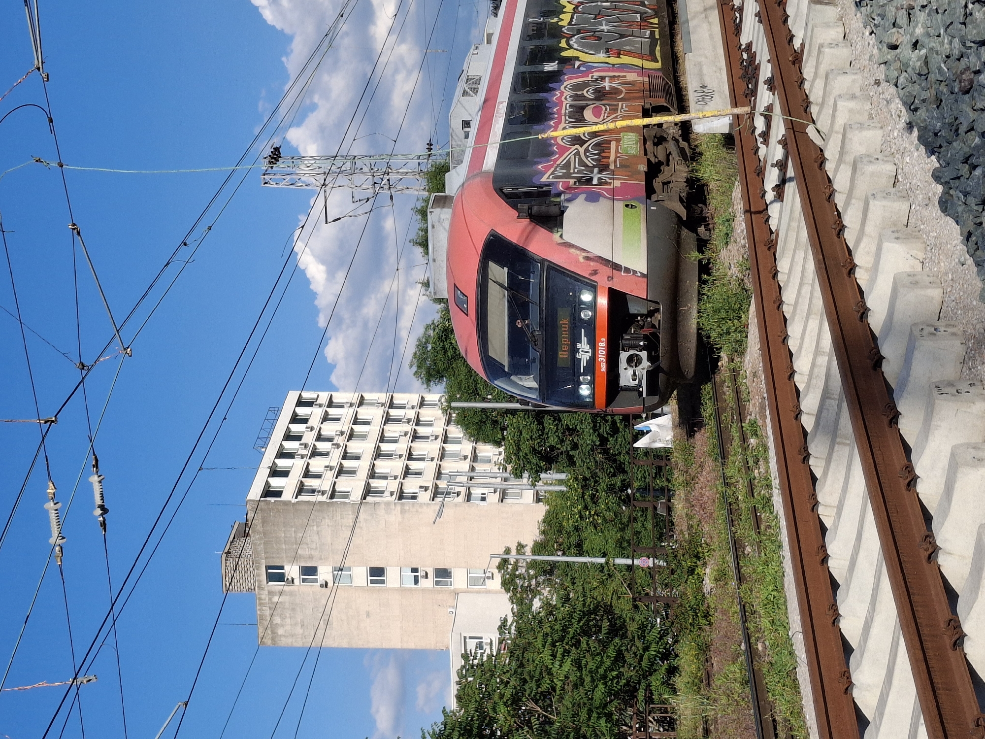 A horizontally oriented, medium-distance shot presents a red and white passenger train, heavily adorned with graffiti, on a track alongside a building. Above, electrical lines crisscross against a partially cloudy sky.
The train occupies the upper right quadrant of the frame. Rust-colored tracks lie adjacent to the train, with the roadbed running parallel to the tracks.
The building is a tall, multi-story structure featuring numerous windows. The sky is blue with scattered white clouds.
The location is in the city of Unknown, Unknown.
