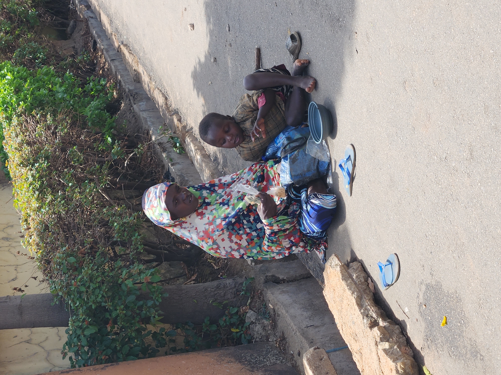 A woman wearing a colorful patterned headscarf and dress sits on the side of a paved road in Jos, Nigeria. She is counting money. Beside her is a young boy with bare feet sitting on the pavement next to a collection bowl. One of the boy's sandals lies nearby on the road. A stick rests near the boy. A second sandal is also visible on the road. The road is bordered by a curb and vegetation.