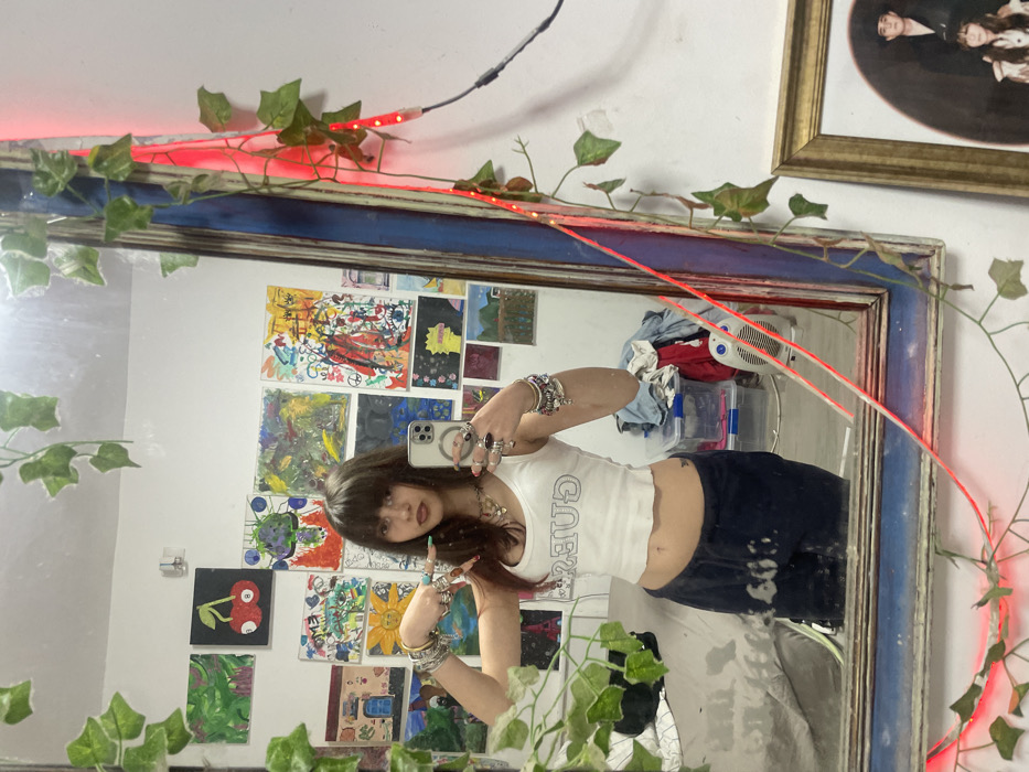A mirrored selfie featuring a young woman with dark hair and bangs. She's wearing a white tank top and dark pants, posing with both hands displaying numerous bracelets and rings. The mirror, adorned with faux ivy and red LED lights, reflects a wall covered in small, colorful paintings. Behind her reflection, glimpses of laundry and a fan are visible. A framed picture hangs on the wall above the mirror.