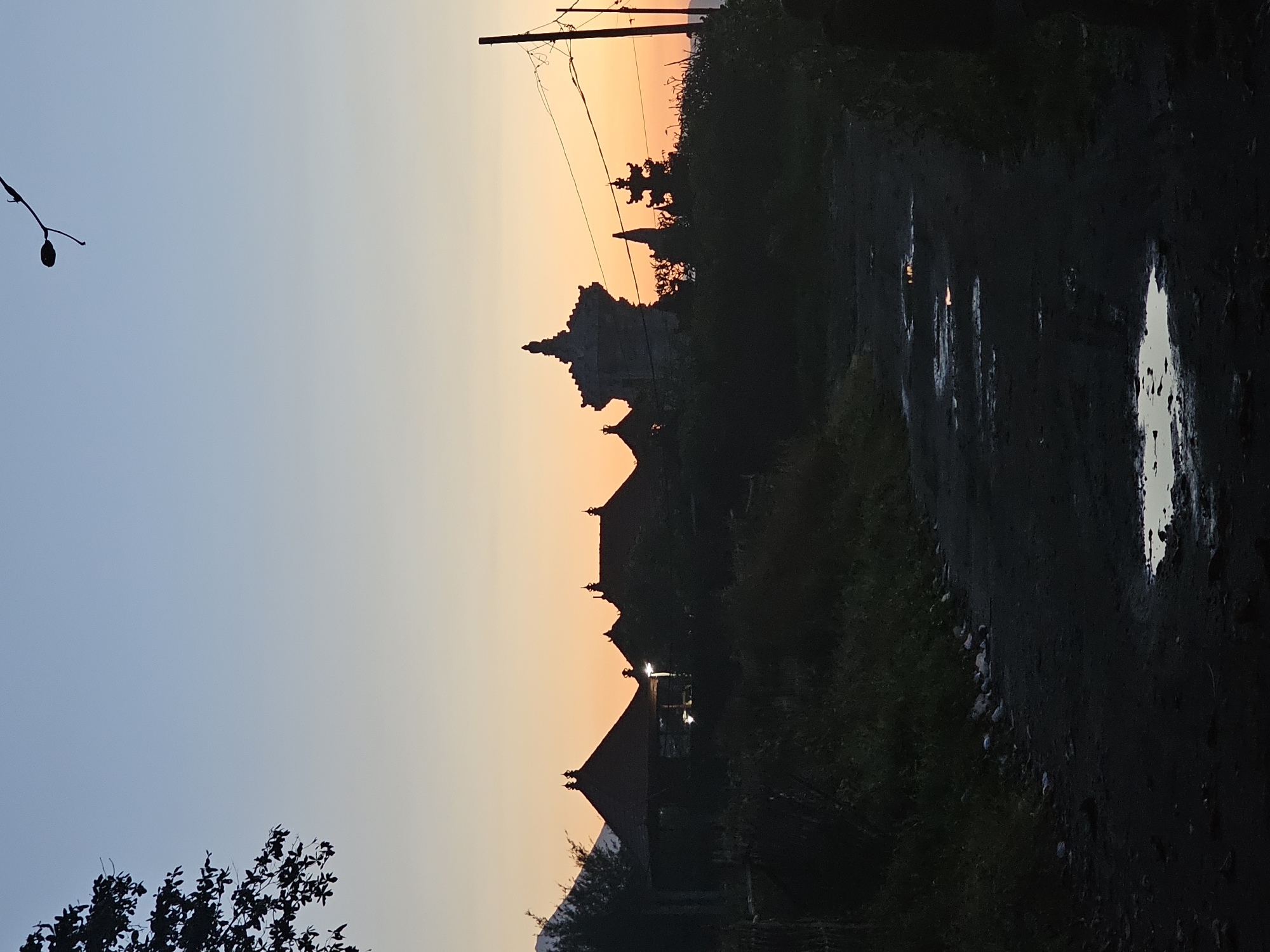 The image depicts a rural scene near Denpasar, Indonesia, during twilight. A dirt road with large puddles reflects the remaining light. The silhouettes of houses, buildings, and trees stand against a sky graded from light blue to orange near the horizon. A utility pole with wires cuts across the upper part of the scene.