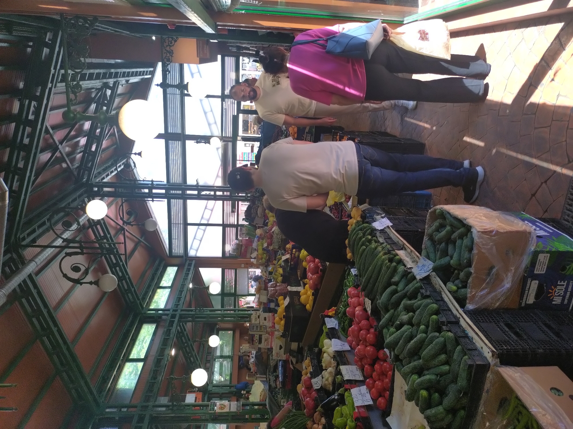 The photograph shows a brightly lit interior space filled with produce, appearing to be the Stara Zagora Central Market Hall in Bulgaria. The high, arched ceiling is made of metal supports and glass panels, casting long shadows across the floor. Rows of stalls display a variety of fresh fruits and vegetables, including tomatoes, cucumbers, and lemons. Several people are visible, including a man with a beard in a light-colored shirt and a woman wearing a pink shirt with a blue purse. A man in a light-colored shirt and a black baseball cap walks along the stall, near cucumbers packed in a box. A mix of customers browse the market, creating an atmosphere of a busy marketplace.