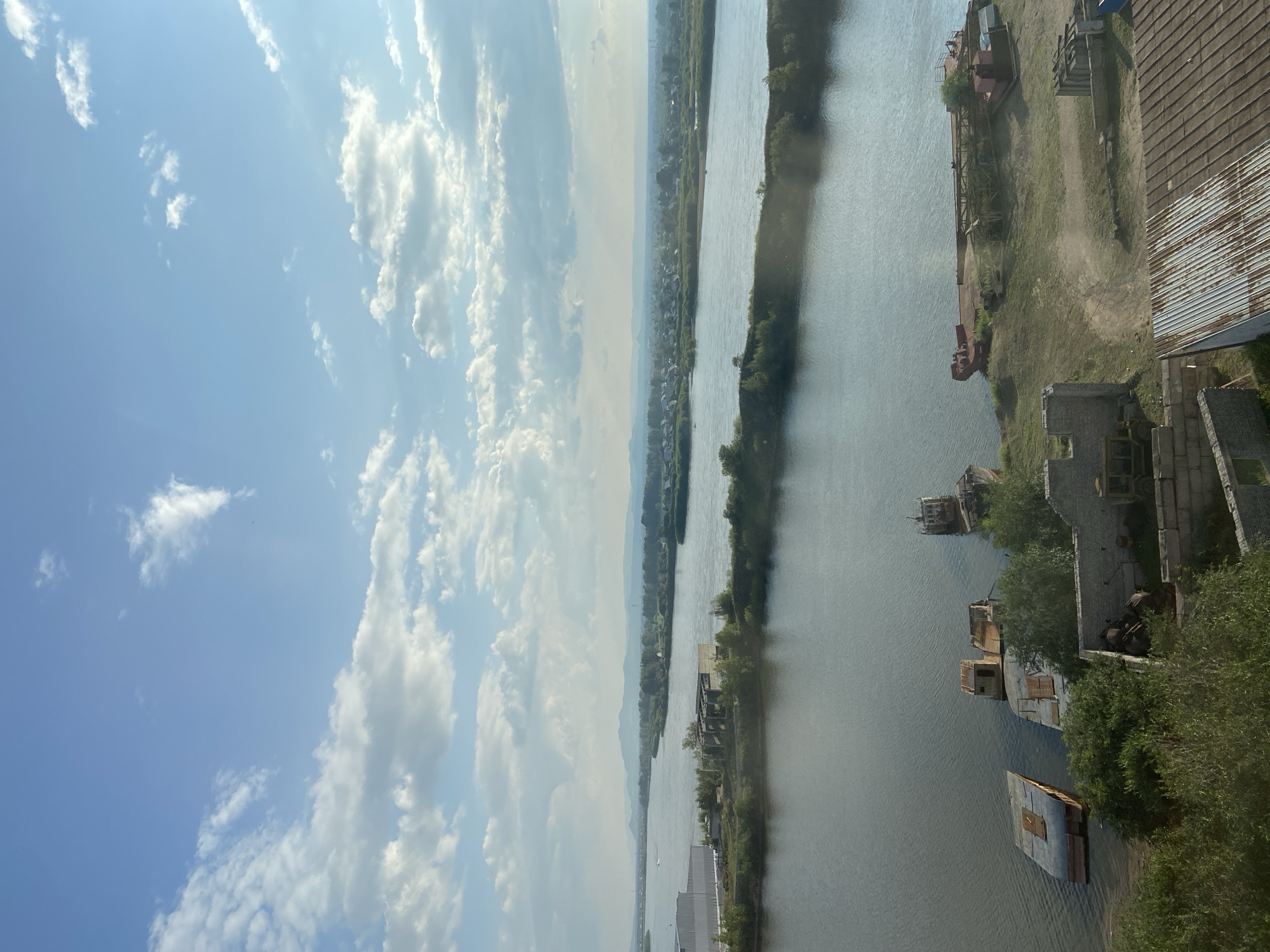 The image captures a wide river, likely the Uda River, flowing through a landscape that suggests the outskirts of Ulan-Ude, Russia. The water is calm and reflects the sky, which is a mix of blue with scattered white clouds.
In the foreground, on the right bank, there is evidence of industrial activity or an abandoned area. Several derelict barges or pontoons are partially submerged or grounded along the shore. Some of these appear to be made of metal and show signs of rust and disrepair. Further inland on this bank, there are the remains of brick structures, possibly old buildings or foundations, and a large crane-like structure. A corrugated metal roof is also visible.
On the left bank, a large, modern building complex with a white facade is present, suggesting active industrial or commercial use. Beyond this, the river widens, and across its expanse, a low-lying town or village is visible, characterized by numerous small houses and buildings nestled amongst green vegetation. The background features rolling hills or mountains under a hazy sky. The overall impression is one of a functional river system adjacent to both industrial and residential areas, with a sense of history conveyed by the abandoned vessels and structures.