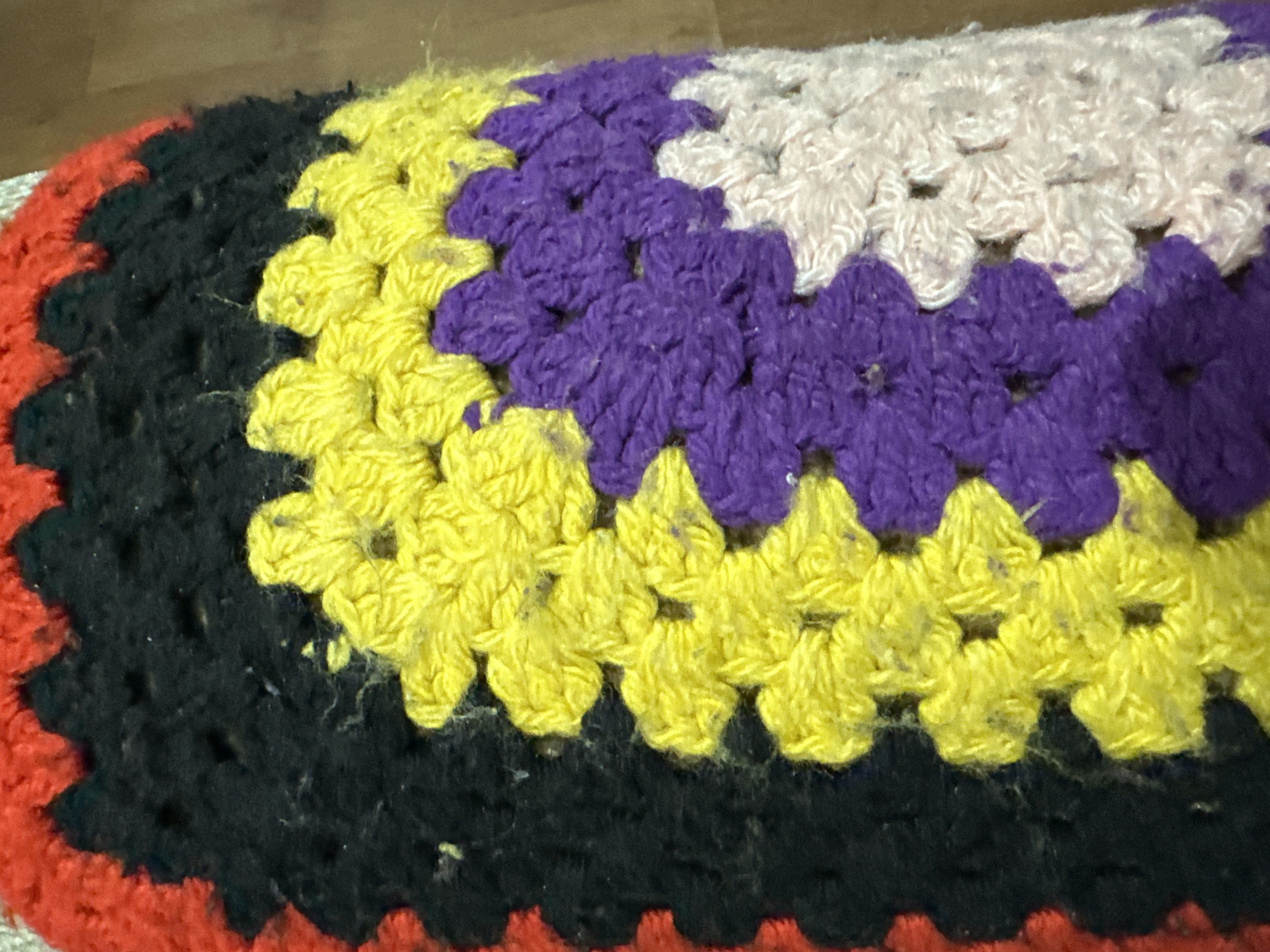 The image shows a crocheted rug or mat. The color scheme consists of concentric bands: starting from the edge, there's a red band, followed by black, then yellow, purple, and finally a light pink or off-white center. The crochet work is visible as individual stitches, giving the rug a textured appearance. The location is Unknown, Unknown.
