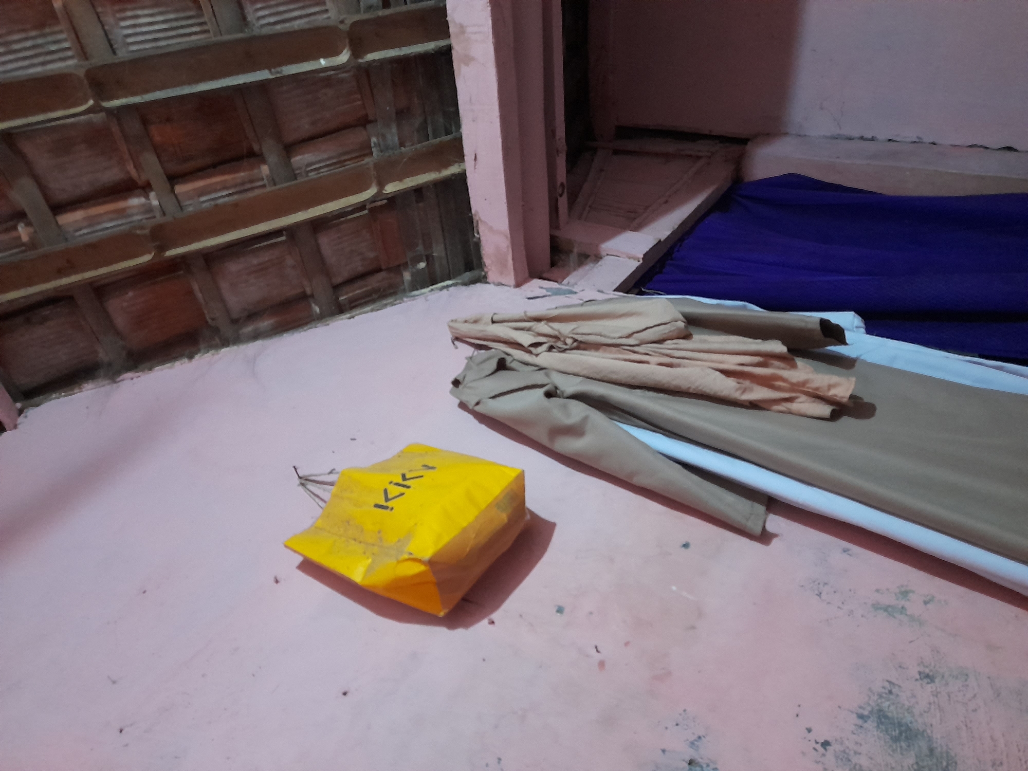 The image shows a humble room with a pink, textured wall. On the wall, a bright yellow shopping bag with the text "KKI" printed on it hangs from a hook. Below the bag, several pieces of clothing are draped over something unseen. A tan long coat and a light blue garment are visible, along with a lighter tan fabric draped over them. To the right, a dark purple fabric hangs, obscuring a doorway or opening. Above, a wooden ceiling with exposed beams and terracotta tiles is visible. The scene suggests a residential interior in a potentially rural or less developed area, possibly Pulokulon, Indonesia, given the context. There are no people visible in the image.