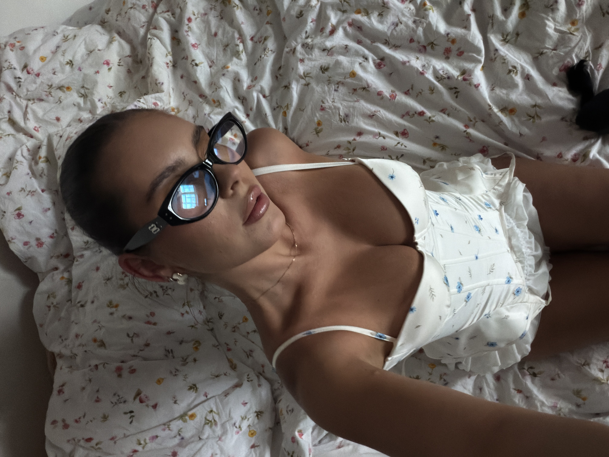 A woman lies on a floral-patterned bed. She is wearing black-framed glasses and a white floral-patterned corset top. She has a gold necklace and is wearing earrings. A black object is visible in the upper right corner of the image. There is no indication that the image was taken in the city of Krasnoyarsk, Russia.