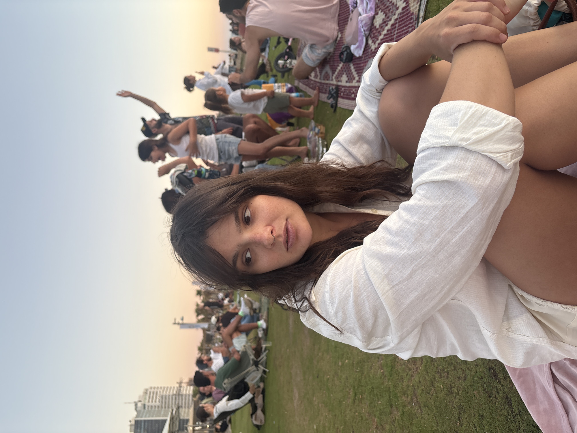 The image shows a young woman in the foreground sitting on a picnic blanket on a grassy area. She is wearing a white linen shirt and appears to be looking off to the side. In the background, a crowd of people are gathered, some sitting on chairs and others standing. The setting appears to be a public park or beach area in Tel Aviv, Israel, with buildings and palm trees visible in the distance. The sky suggests it is late afternoon or early evening.