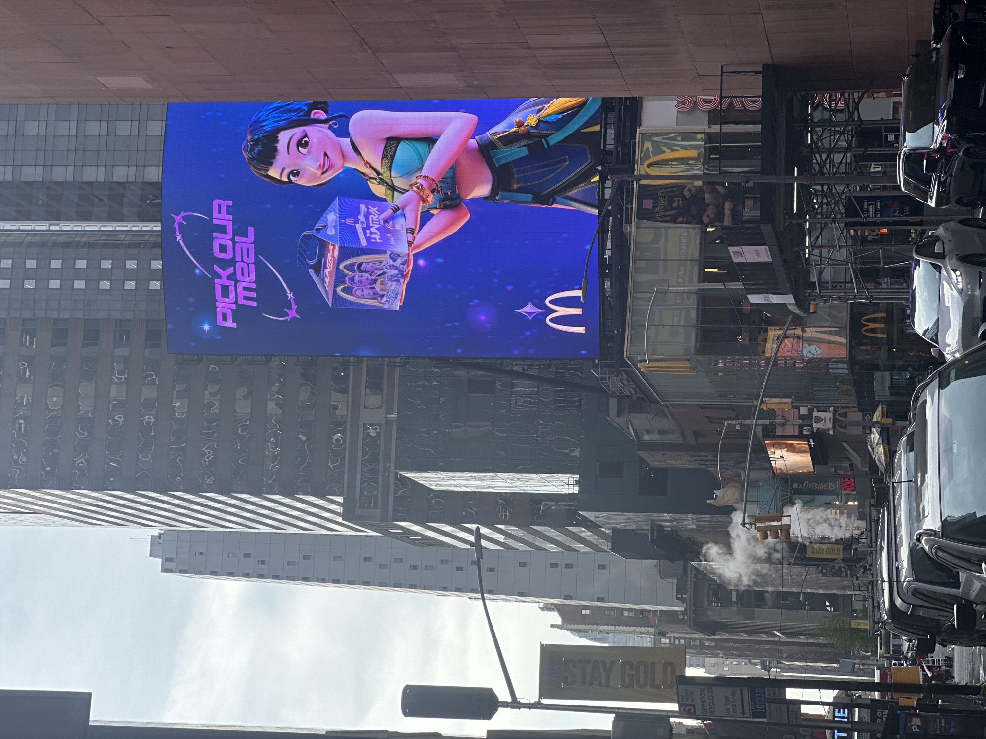 The image depicts a scene in New York City, USA, featuring a large digital billboard advertising a McDonald's meal, "PICK OUR MEAL". The billboard displays an animated character holding a "The Huntrax" meal box, set against a blue background with the McDonald's logo. The surrounding environment includes typical NYC street elements such as buildings with reflective windows, street signs (including "STAY GOLD" and parking signs), steam rising from the street, traffic lights, and a row of parked vehicles including cars and vans. There are also signs for "Outsiders" visible on the buildings. The presence of these elements suggests a busy urban environment, typical of the Times Square area in Manhattan.