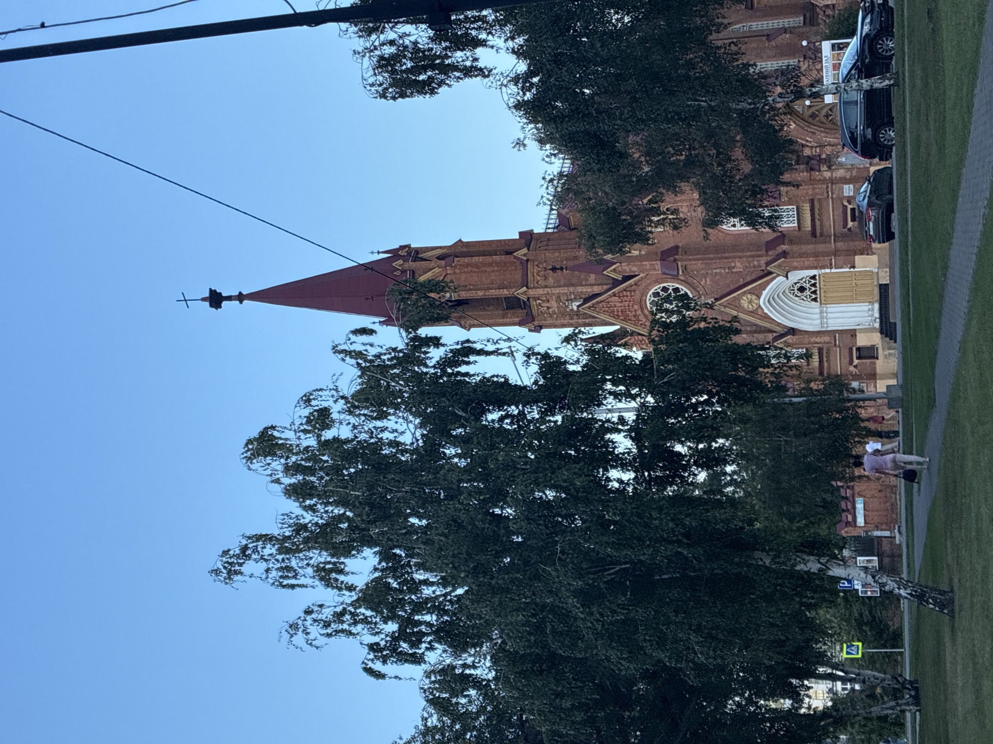 The image shows the Roman Catholic Cathedral of the Immaculate Conception of the Virgin Mary in Irkutsk, Russia. The church is a prominent brick building with a tall red spire topped by a cross. In the foreground, several tall, leafy trees partially obscure the view of the church. A paved walkway runs in front of the church, and a grassy area is visible to the left. Two cars are parked near the entrance of the church. A few people are walking on the sidewalk and in the distance. The sky is a clear blue, indicating daylight. The overall scene is one of urban architecture and nature coexisting.