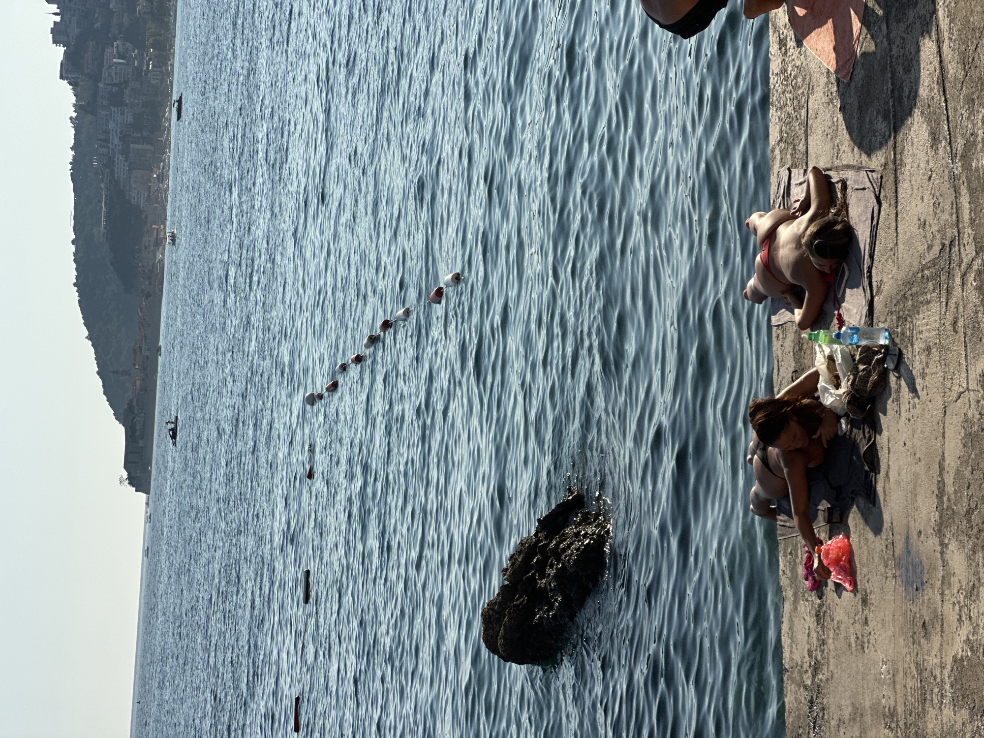 Two women are sunbathing on a concrete pier next to the Adriatic Sea in Budva, Montenegro. The woman on the left is lying on her stomach, wearing a dark bikini, and the woman on the right is lying on her back, wearing a red bikini. Both are on towels. A string of buoys is visible in the water. In the background, a rocky outcrop emerges from the sea, and a town with buildings and hills is visible on the coastline. The sky is clear and blue.