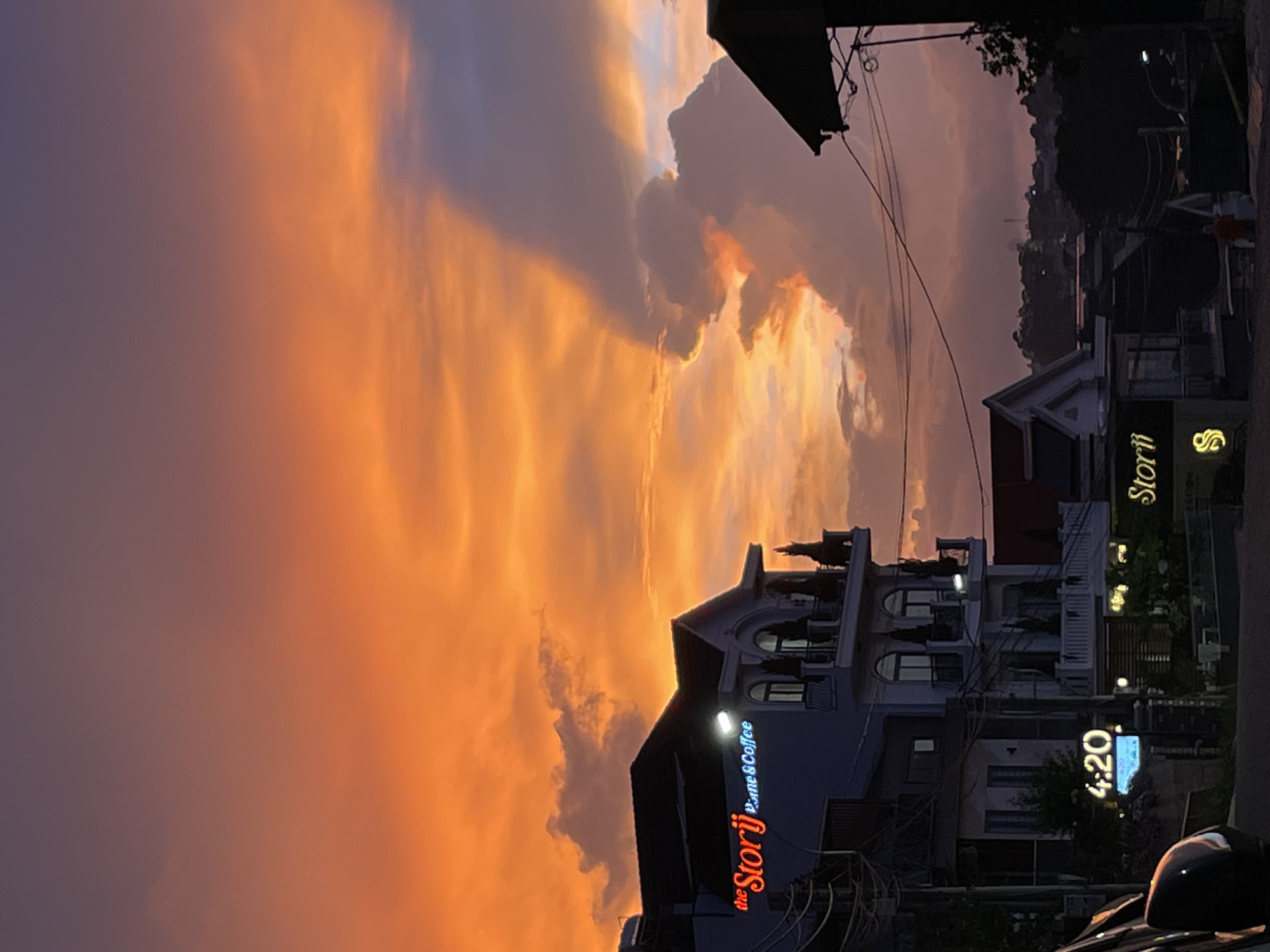 The image shows a street scene at dusk, with a vibrant orange and purple sky filled with dramatic clouds. In the foreground, on the right side, is a building with several stories and balconies. The building is illuminated with neon signs. One sign reads "the Storij Home & Coffee" in red and blue neon. Another sign on a different building, to the right, is not entirely legible but appears to have a stylized "S" logo and the word "Storii". Further to the right, a digital clock displays "4:20".
The setting appears to be a town or city, with other buildings visible in the background. Trees and power lines are also present, suggesting an urban or suburban environment. The time of day is clearly sunset or twilight, as indicated by the colorful sky and the artificial lighting from the signs. The overall mood is atmospheric, with the dramatic sky dominating the scene. The presence of businesses like a "Home & Coffee" suggests a commercial area. The location context provided is Lâm Viên - Đà Lạt, Vietnam. No people are clearly visible and engaged in any specific activity, though a blurred figure in red can be seen in the background on the far right.