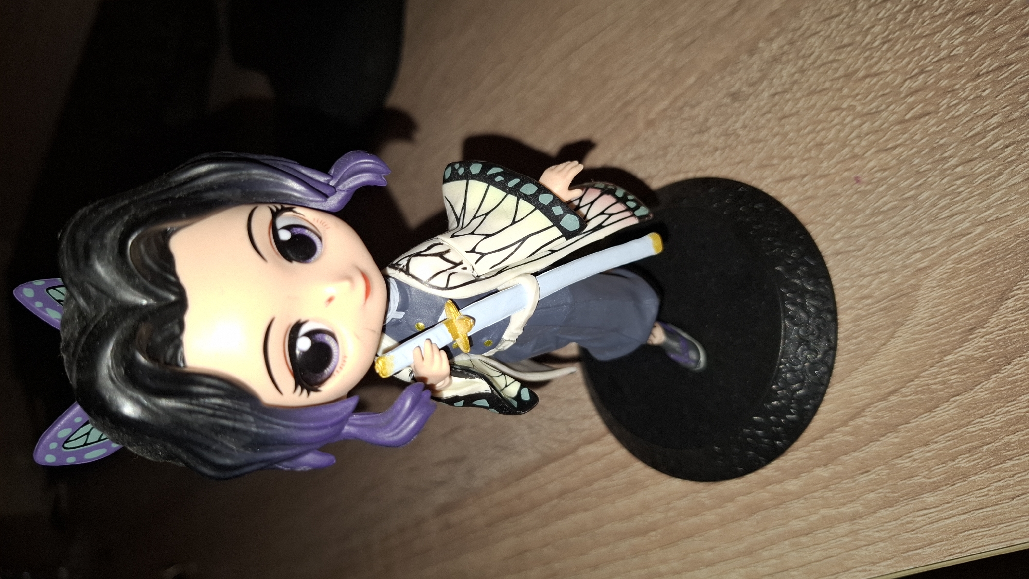 The image depicts a Shinobu Kocho figurine from the anime series Demon Slayer. The figurine is positioned on a dark base with textured edges. The figurine itself has a cartoonish, chibi-style appearance. She has black hair with purple tips and butterfly-shaped hair ornaments. Her outfit consists of a blue and white uniform with a butterfly-patterned haori. She is holding a sword with a yellow hilt and a blue blade. The figurine appears to be placed on a wooden surface. The background is partially dark, suggesting a dimly lit environment. No information from the image suggests that it was taken in Sofia, Bulgaria.