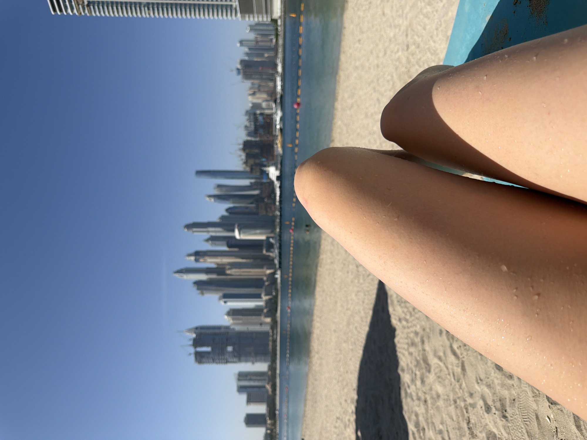 The image shows a close-up of a person's legs with water droplets on the skin, likely after swimming. The legs are resting on a sandy beach. In the background, the skyline of Dubai, United Arab Emirates, is visible, indicating the location. The water from the Arabian Gulf runs adjacent to the beach, with a line of buoys visible in the water. The sky is clear and blue, suggesting a sunny day. The skyscrapers of Dubai's Marina district are the dominant visual element of the background, identifying the general location.