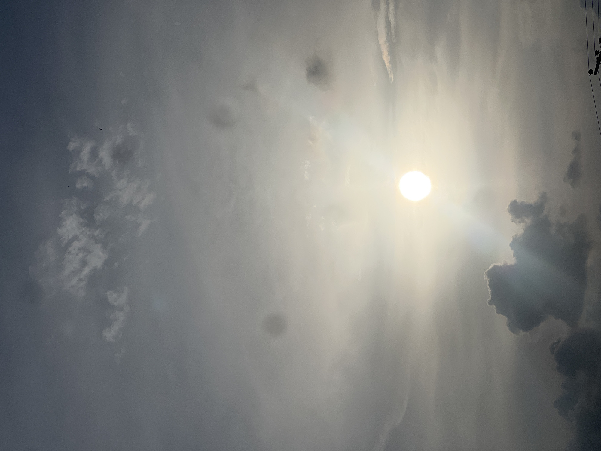 This is a photograph of a cloudy sky with the sun visible and slightly obscured by the clouds. The sun is a bright, luminous orb in the center-left of the image. There are various cloud formations in the sky, ranging from light, wispy clouds to darker, more dense formations. In the lower right corner of the image, there are electrical wires and a pole visible, suggesting an urban or semi-urban environment. There are no people or animals visible in the frame. The overall impression is that of a partly cloudy day with the sun attempting to break through. There is no specific indication of the location being Kporva, Nigeria, in the image itself.