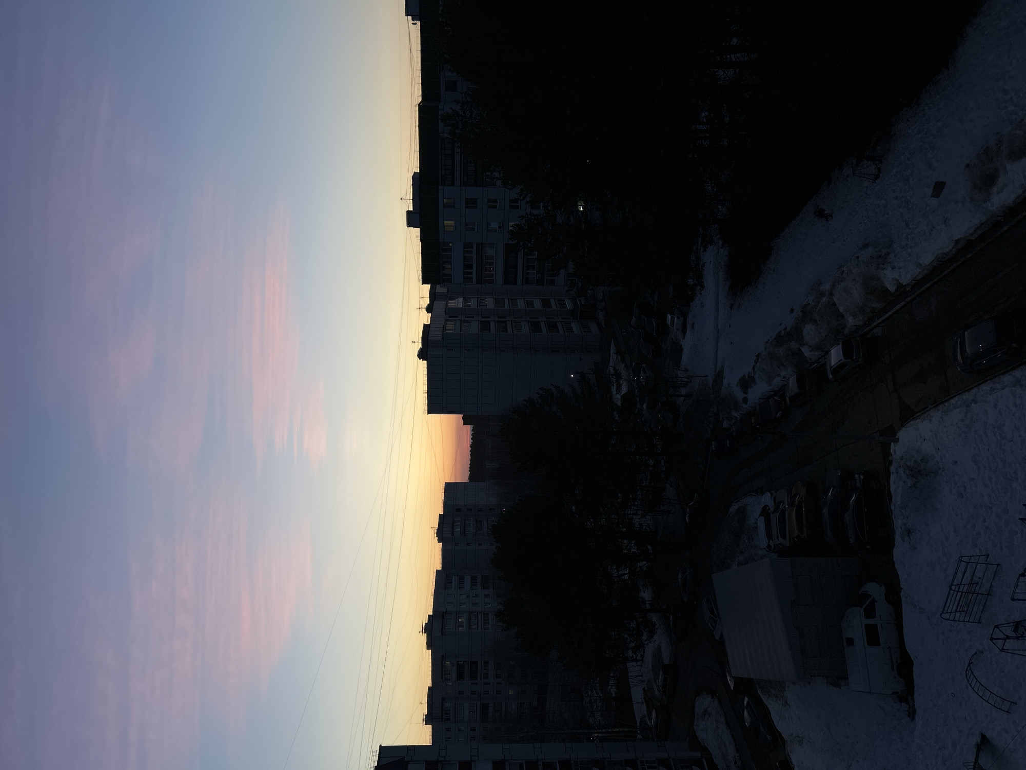The image depicts an evening scene in what appears to be a residential area of Chaikovsky, Russia. The sky transitions from shades of blue and pink near the horizon to darker hues higher up. The lower portion of the image is obscured by buildings, trees, and snow-covered ground. Some cars are visible in a parking area, and some children's play equipment is also apparent, suggesting a public space in a residential environment. There are no visible people in the scene.