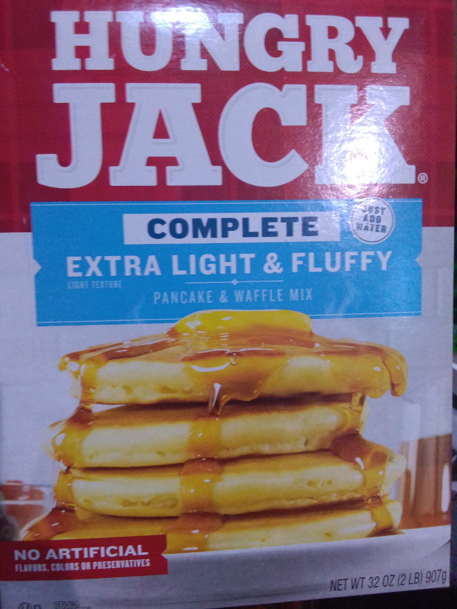 The image is a close-up of a box of Hungry Jack pancake and waffle mix. The box features prominent branding with "HUNGRY JACK" written in large white letters against a red background. Below this, a blue banner contains the text "COMPLETE EXTRA LIGHT & FLUFFY PANCAKE & WAFFLE MIX." A small circular graphic with "JUST ADD WATER" is also visible on the blue banner. The bottom portion of the box shows a stack of syrup-drizzled pancakes, with a red banner below that reads "NO ARTIFICIAL FLAVORS, COLORS OR PRESERVATIVES." The text "NET WT 32 OZ (2 LB) 907g" is visible at the bottom right corner of the box. The location of Howayi, Nigeria is not depicted in this image.