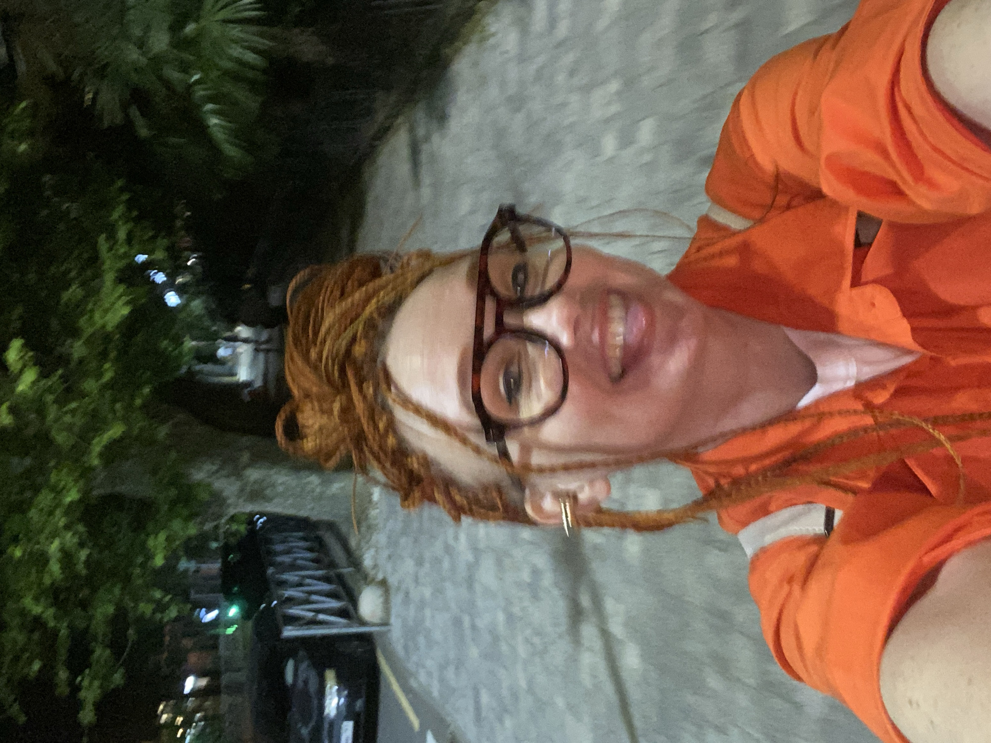 A person with ginger-colored dreadlocks, wearing glasses and an orange jacket, takes a selfie while walking on a cobblestone sidewalk at night. Trees and a fence line the street, with cars parked on the left side. The scene appears to be in a city, possibly Sochi, Russia, given the context. The person is smiling and sticking out their tongue.