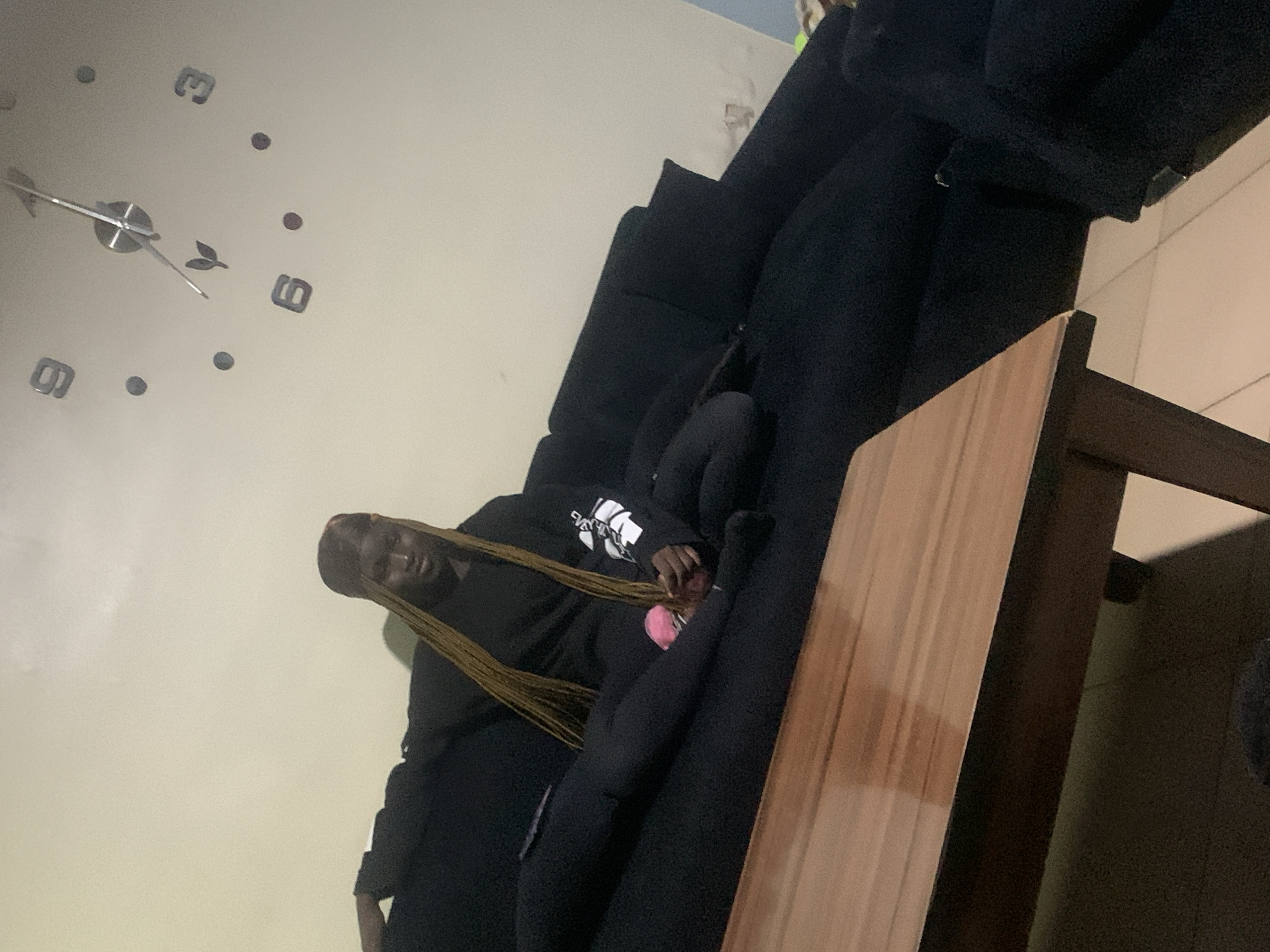 A woman with long, blonde braided hair and a black headscarf sits on a large, dark sectional sofa in an indoor living room. She is wearing a black sweatshirt with white lettering on the right sleeve and black leggings. Her right leg is bent, with her foot resting on the sofa, and her left leg is extended. Her hands are resting on her lap. In the background, on a light-colored wall, is a large, modern clock with Roman numerals and metallic hands. In the foreground, a portion of a wooden coffee table is visible. The room appears dimly lit.