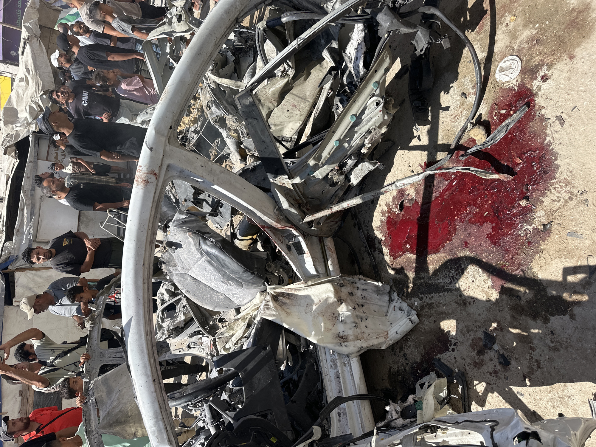 The image depicts the aftermath of an incident in Gaza City, Palestinian Territories. In the foreground, a severely damaged white vehicle is visible, its interior exposed and mangled. Fragments of metal and debris are scattered around the car. A significant pool of red liquid, consistent with blood, is present on the ground near the vehicle. Several individuals, appearing to be civilians and possibly first responders, are gathered around the scene, observing the damage. Some are looking at the car, while others are engaged in conversation or taking photographs. The environment appears to be an urban setting, with damaged structures and tents visible in the background, suggesting a context of conflict or disaster.