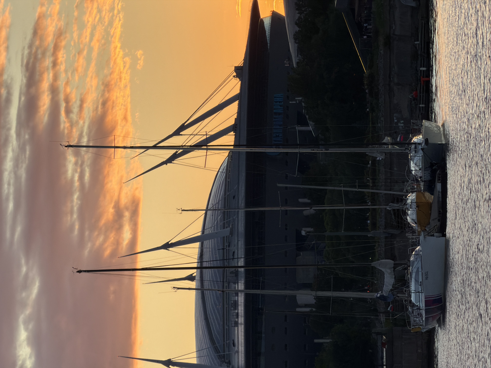 The image captures a serene waterfront scene at sunset in Saint Petersburg, Russia. The sky is a canvas of warm oranges and soft yellows, with wispy clouds adding texture. Dominating the background is the distinctive architecture of the Gazprom Arena, its futuristic design silhouetted against the colorful sky. Several sailboats are moored in the foreground, their masts reaching upwards like elegant fingers. The calm water reflects the warm hues of the sunset, creating a peaceful and picturesque atmosphere. The time of day is clearly sunset, indicated by the vibrant colors in the sky. The location is identified by the presence of the Gazprom Arena and the general waterfront setting typical of Saint Petersburg. The scene evokes a sense of tranquility and beauty. Visible text on one of the boats reads "PAV-2002".