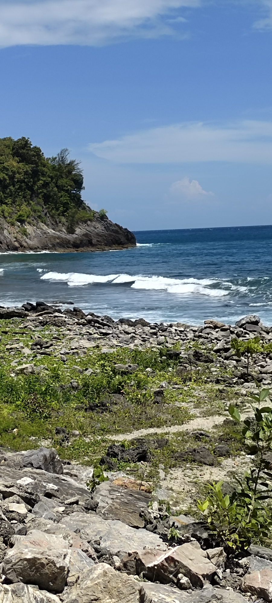 This is a vertical, eye-level shot of a rocky coastline in Naga Umbang, Indonesia. The foreground is filled with a jumble of grey and tan rocks of various sizes, interspersed with patches of green vegetation. A narrow, sandy path winds through the rocks in the middle ground. Beyond the rocks, a body of dark blue water stretches out to the horizon, with white waves crashing near the shore. On the left side of the image, a rocky, tree-covered cliff juts out into the sea. The sky above is a clear, bright blue with a few wispy white clouds. No people or animals are visible in the image.