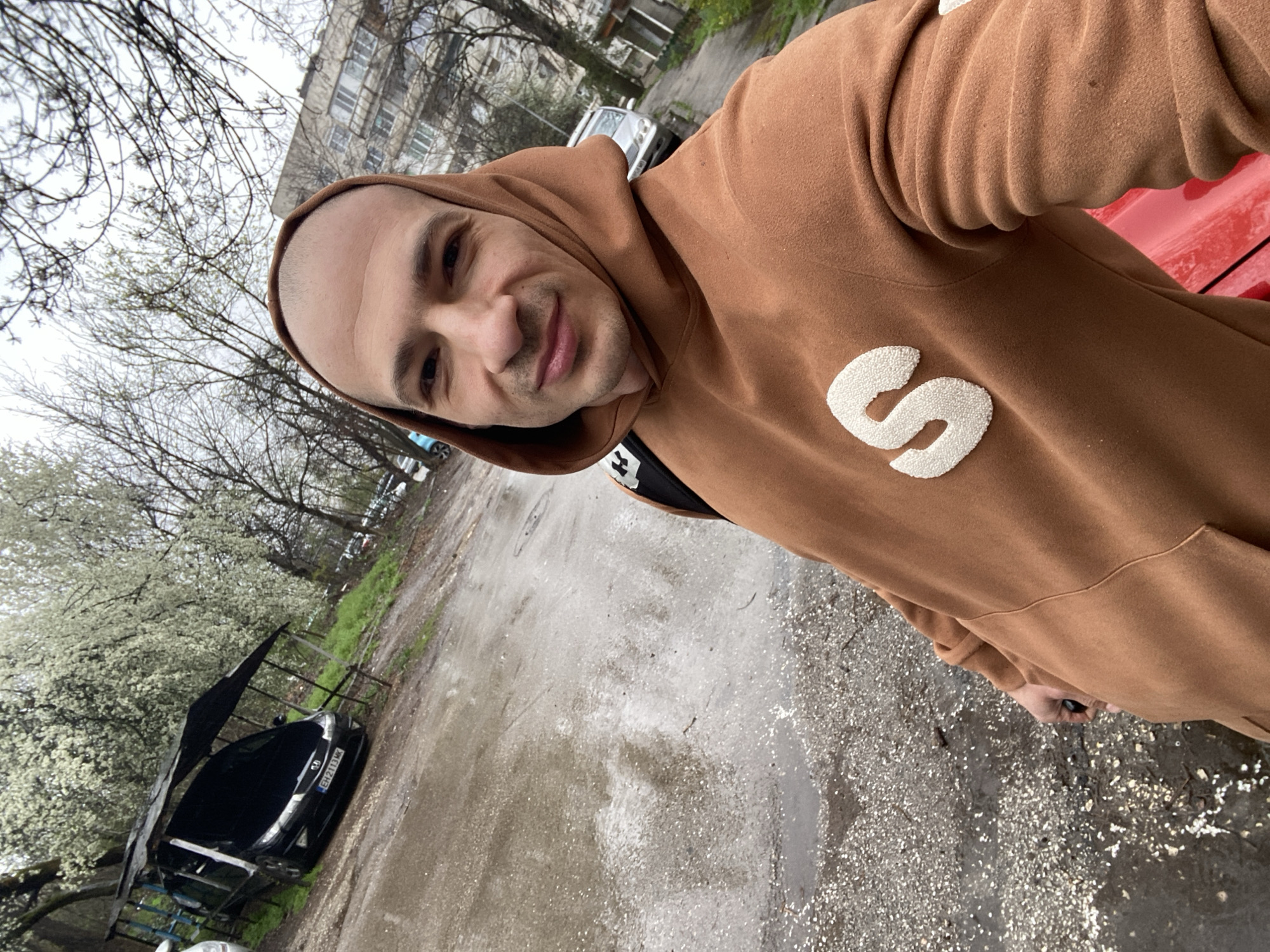 A man is taking a selfie. He is wearing a brown hoodie with a white "S" on the chest. He appears to be standing on a wet, cracked asphalt surface. Behind him is a car parked under a makeshift shelter, with trees and a multi-story building in the background. The car's license plate reads "EH7213MK". Based on this license plate, the image is taken in the city of Pleven, Bulgaria.