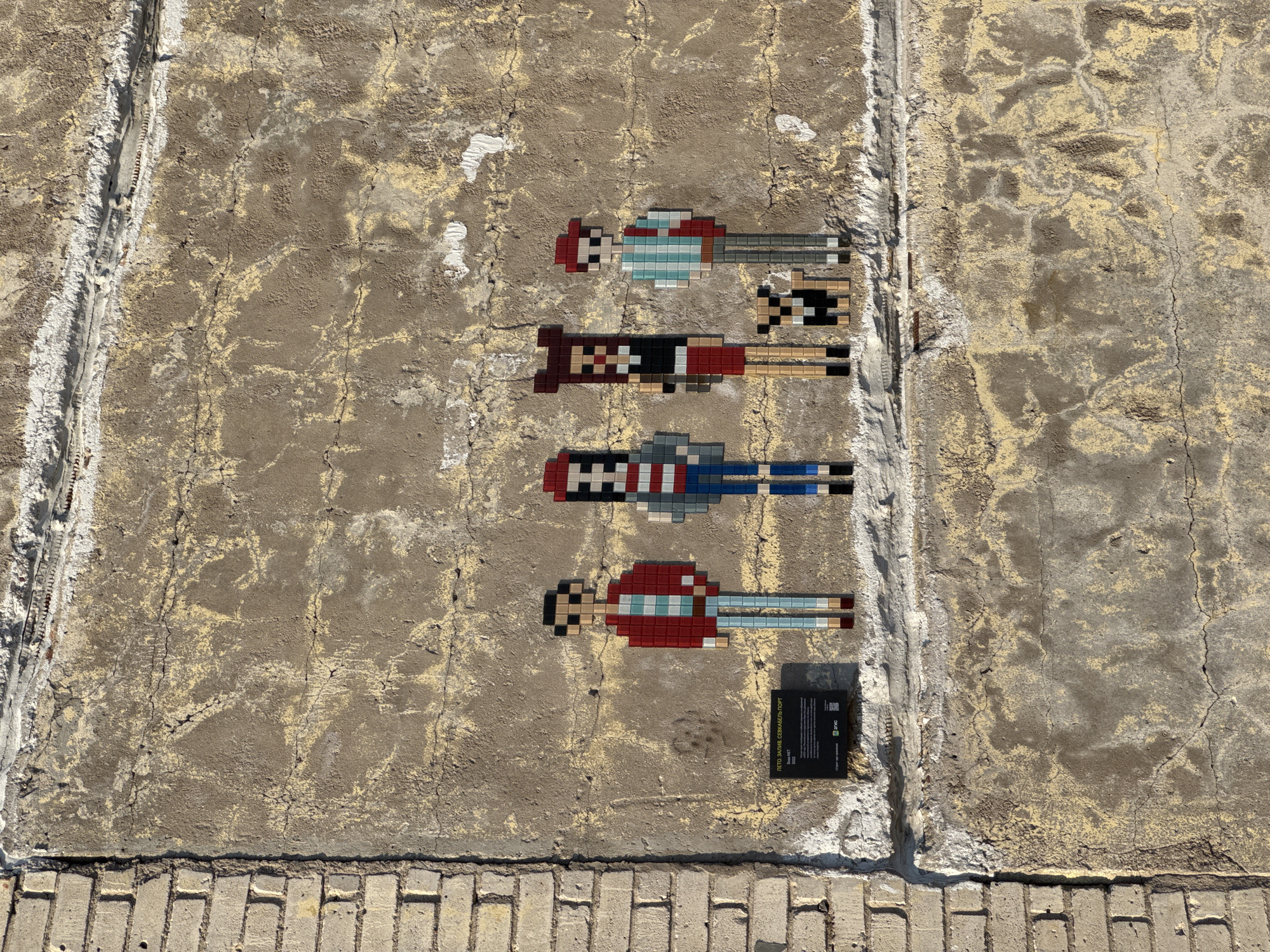 The image displays an outdoor wall adorned with four pixel-art mosaic figures. The wall itself is a weathered, textured concrete or stucco surface, characterized by patches of light yellow discoloration, numerous cracks, and signs of general wear. A rough vertical seam or groove runs down the left side of the wall, and a horizontal line of light-colored bricks or pavers forms the bottom edge of the frame.
The four mosaic figures are stylized, humanoid shapes composed of small, square tiles in various colors, evoking an 8-bit video game aesthetic. They are arranged vertically, lying horizontally on the wall, with their "heads" generally facing towards the left side of the image. Each figure exhibits a distinct color scheme: the topmost has a red hat, a light blue and white striped shirt, and grey pants; the second features a dark brown and red top with light brown pants and black shoes; the third wears a grey top with red and white accents and blue pants; and the bottommost is depicted with a red top, a light blue and white striped mid-section, and light blue pants.
To the bottom right of the lowest mosaic, a small, black rectangular plaque is affixed to the wall. The plaque contains text, predominantly in Cyrillic, and a QR code. The legible Cyrillic text at the top reads "ЛЕТО, ЗАГИНИ, СЕВКАБЕЛЬ ПОРТ" ("Summer, Bend, Sevkabel Port"), which refers to a known cultural space in Saint Petersburg, Russia. Below this, "Invader NET" and "2022" are visible, likely indicating the artist or series name and the year of creation. A small logo, possibly "SYNO", is also present.
The scene is brightly illuminated by natural light, suggesting daytime, with harsh shadows indicating direct sunlight. The overall impression is that of an urban street art installation or a public artwork set against a worn, industrial-like backdrop, consistent with its location at Sevkabel Port in Saint Petersburg, Russia.