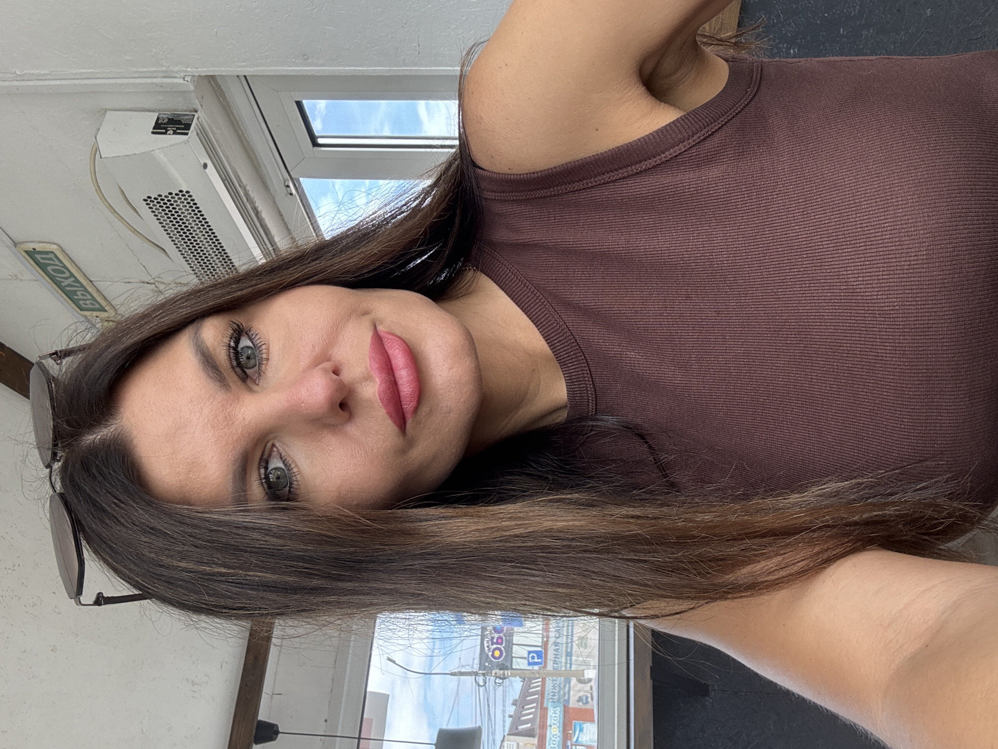 A woman with long, dark brown hair and green eyes is pictured in a selfie taken indoors, likely in Chaikovsky, Russia, given the presence of a sign that reads "ВЫХОД" (EXIT) in Russian. She is wearing a brown, ribbed, sleeveless top. Sunglasses are perched on top of her head. The background includes a white air conditioning unit mounted on a white wall, a window with a view of buildings and power lines, and a wooden beam overhead. The lighting suggests it is daytime.