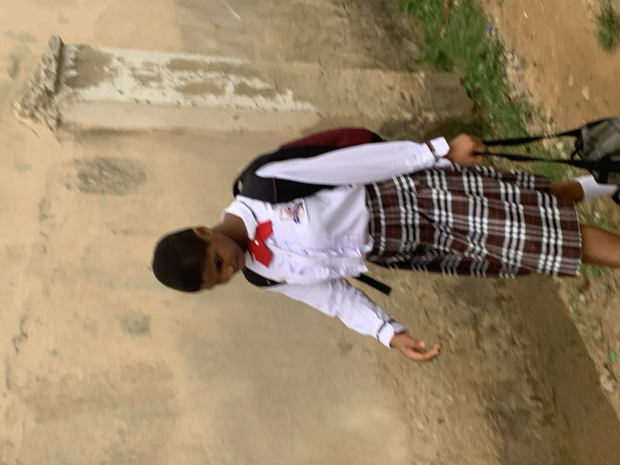 A young person wearing a school uniform consisting of a white button-down shirt with a red bowtie, a plaid skirt, and white socks is walking along a dirt path beside a textured, light-colored wall. The person has short dark hair and is looking downwards. They are carrying a dark backpack on their shoulders and holding a black bag in their right hand. Green vegetation is visible along the bottom right of the image. The scene appears to be outdoors in a seemingly urban or semi-urban environment, possibly in Abuja, Nigeria, given the architectural style of the wall and the commonality of school uniforms.