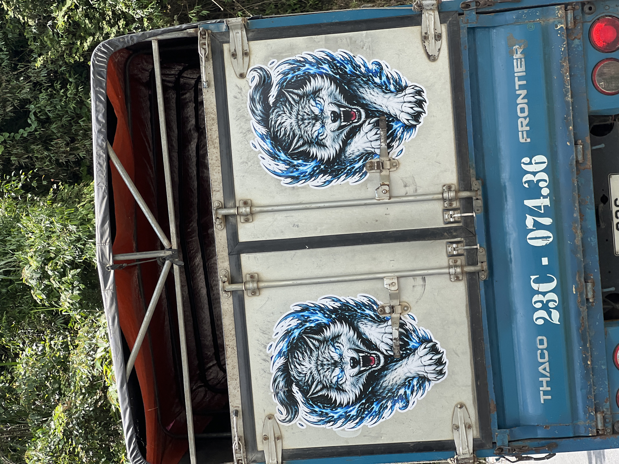 The image shows the rear of a blue truck parked on a paved surface, possibly a road. The truck has a white license plate with Vietnamese characters and numbers, reading "23C - 074.36". The truck also features the brand name "THACO" and "FRONTIER" in white lettering. Two stickers of a fierce-looking wolf with blue flames are affixed to the truck's metal rear doors. The background consists of dense green foliage, indicating a natural environment, possibly a rural area in Quản Bạ Commune, Vietnam, given the context. Sunlight illuminates the scene, casting shadows and highlighting the details of the truck and its surroundings.