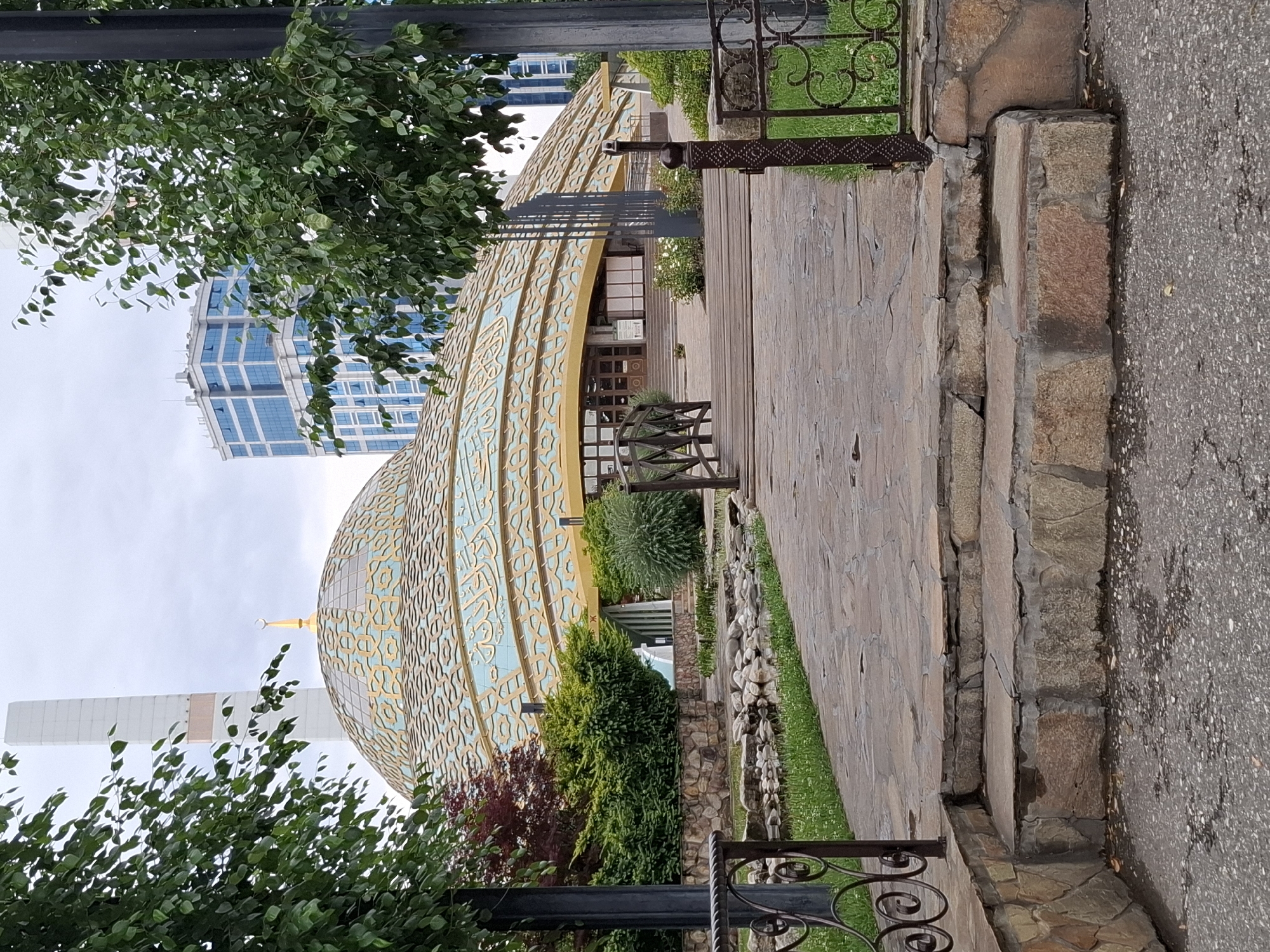 A paved path leads to the Argun Mosque, also known as the Mosque of the First President of the Chechen Republic Akhmad-Khadzhi Kadyrov, in Argun, Russia. The mosque features a large, ornate dome with Arabic inscriptions and a golden crescent moon on top. A tall, white minaret stands to the left of the dome. The path is made of stone slabs and is lined with decorative metal railings and lush green bushes. In the background, a modern blue glass skyscraper rises above the trees. The sky is overcast, suggesting a cloudy day.