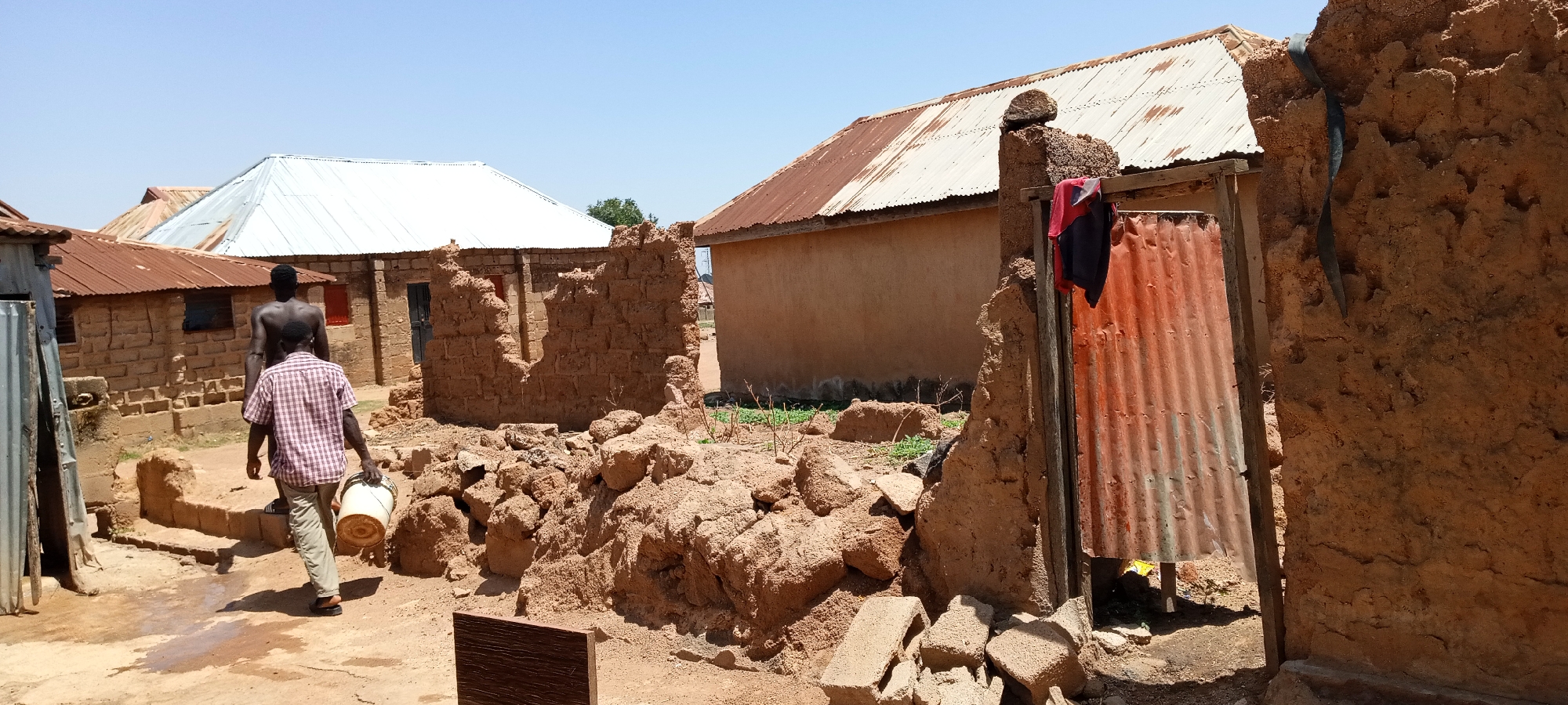 In Jos, Nigeria, the image shows an outdoor scene in what appears to be a residential area with damaged mud structures. Two men are present: one wearing a plaid shirt and khaki pants, carrying a white bucket, and another walking behind him, shirtless. The buildings have corrugated metal roofs, some appearing rusted or old. Debris from collapsed walls is scattered on the ground. The environment is sunny and dusty. A piece of cloth is hanging on a partly collapsed structure.