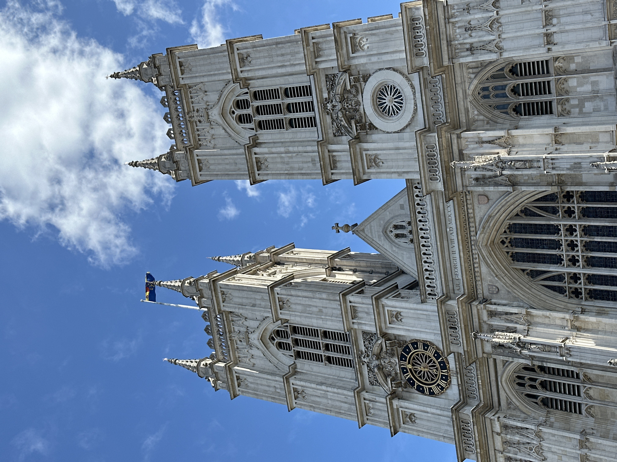 This low-angle shot depicts the towers of Westminster Abbey in London, United Kingdom, against a bright blue sky with scattered clouds. The towers are constructed of light-colored stone and feature intricate Gothic architectural details, including spires, arched windows, and ornate carvings. A clock is visible on one of the towers, and a flag flies from another. The inscription "A.R. GEORGII. II. VIIL A.D. MDCCXX" can be seen on the building. The towers dominate the frame, emphasizing their height and architectural complexity.