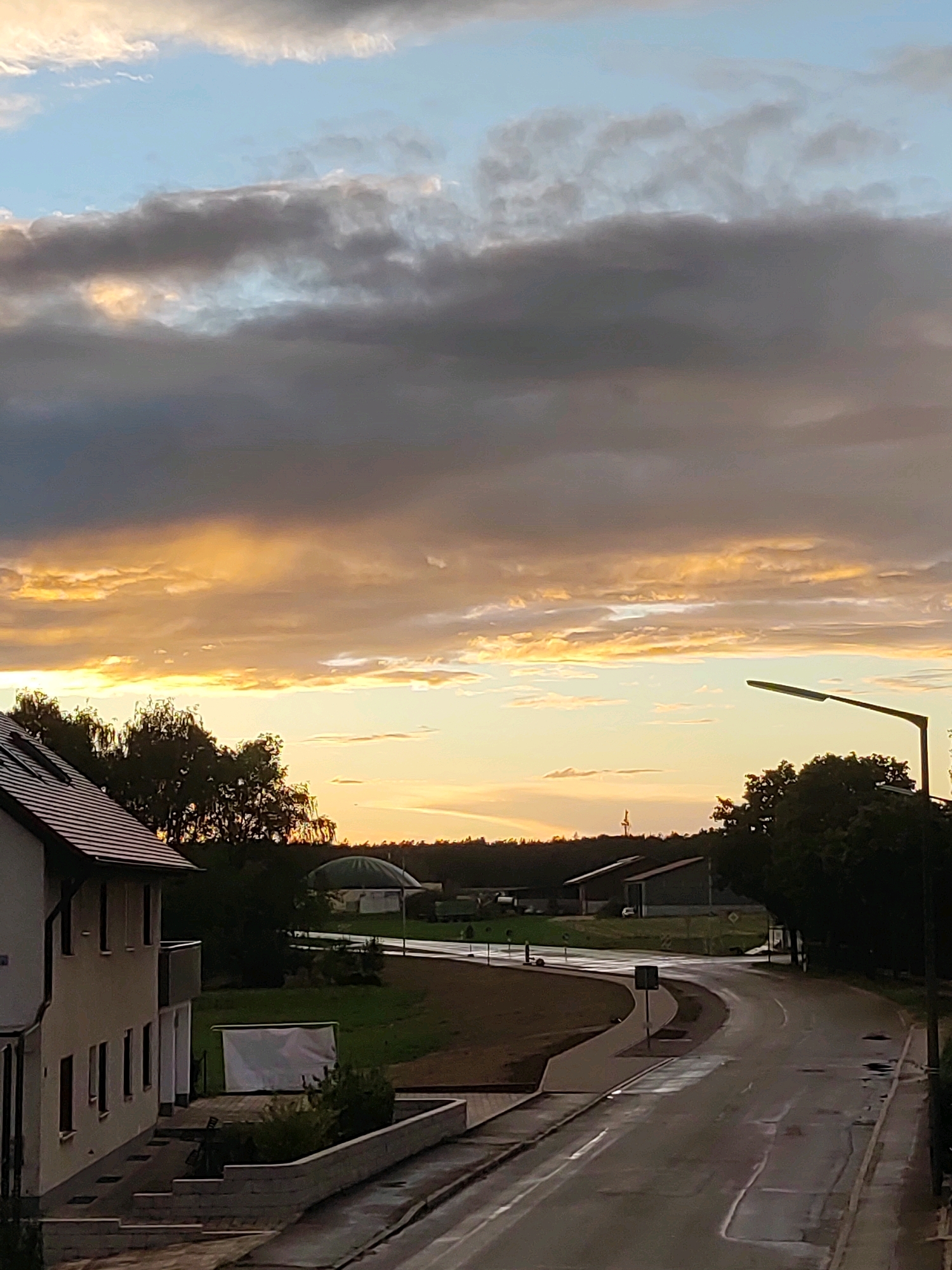 The image depicts a street scene in Burgoberbach, Germany, likely during a sunset. The sky is filled with dramatic clouds, with streaks of golden and orange light breaking through the darker, grayer portions. In the foreground, a modern building with a tiled roof and windows is visible on the left, with a stone wall and steps leading up to it. A paved road curves through the middle ground, wet from recent rain, reflecting the sky's colors. On the right side of the road, a tall street lamp stands illuminated. Beyond the road, there are trees, some farm buildings with distinctive roofs, and a dense line of trees forming the horizon. The overall impression is one of a rural or semi-rural setting on the outskirts of a town, with a dynamic sky as the main focal point.