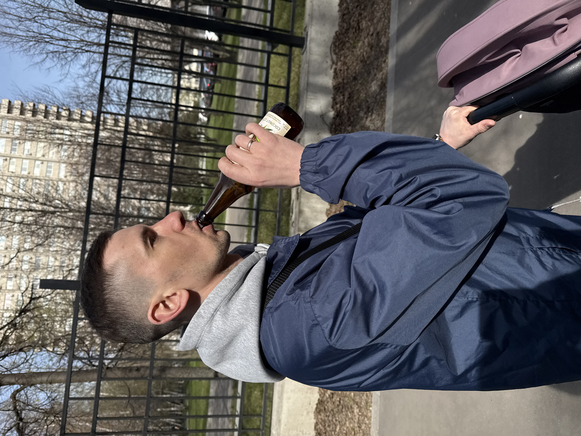 A man is captured in a profile view, walking outdoors on a bright, sunny day in Moscow, Russia. He is actively drinking from a brown glass bottle held in his right hand, while simultaneously pushing a pink-canopied baby carriage with his left hand. He wears a casual outfit comprising a grey hooded sweatshirt beneath a dark blue jacket, and a ring is visible on his left ring finger. The bottle's label is partially visible, showing "COCONUT C..." in white text against a green and white background.
The scene depicts an urban outdoor environment. A black metal fence separates the paved path where the man walks from the background. Visible through the fence is a large, light-colored multi-story building with numerous windows and a distinctive, crenellated roofline. Bare trees are scattered in the background, and the sky above is clear and blue, indicating pleasant weather. The overall activity suggests a casual stroll.
