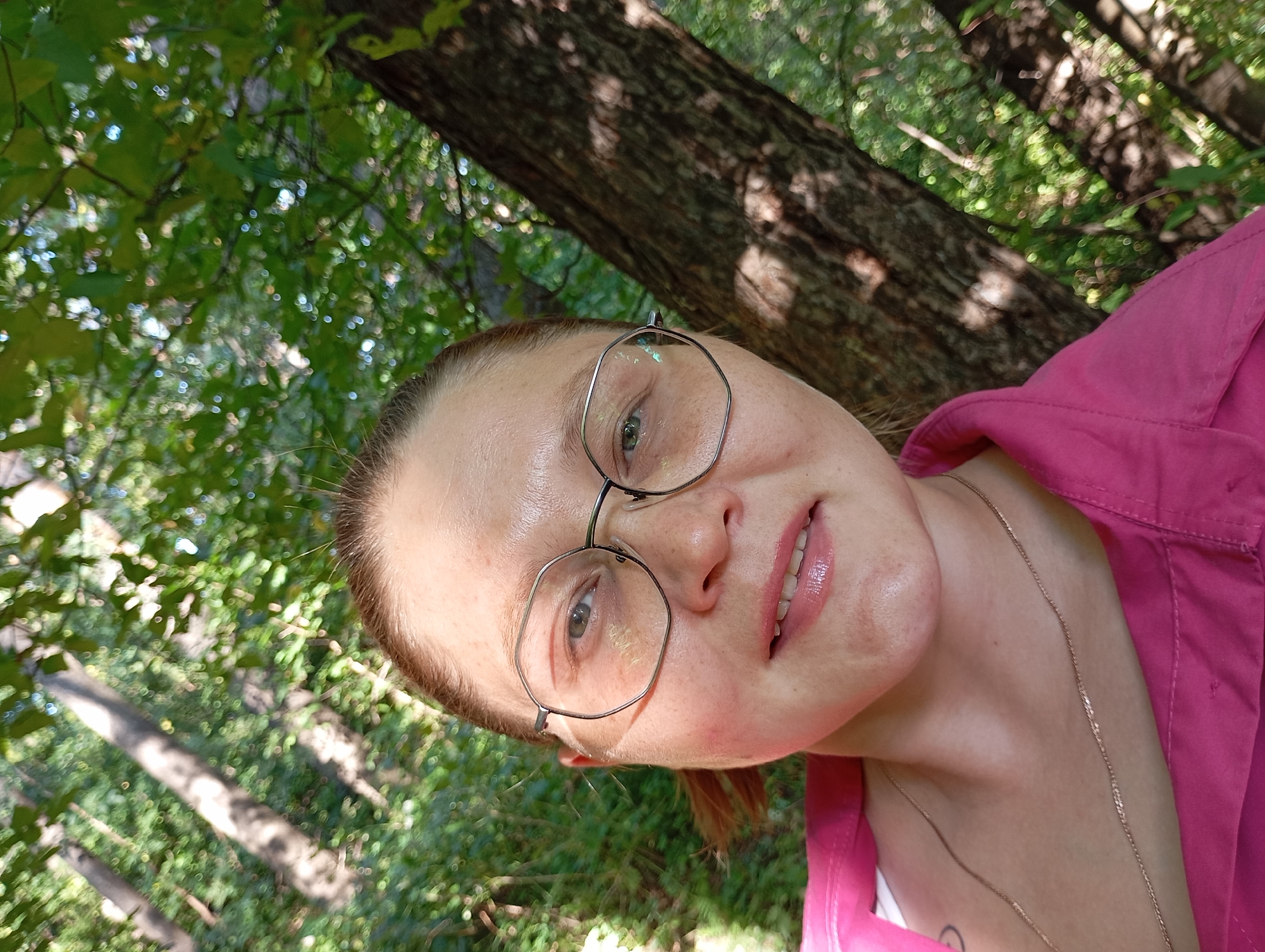 A low-angle, selfie-style shot features a light-skinned woman with short blonde hair, wearing glasses with hexagonal frames and a bright pink button-up shirt. She is outdoors, with a tree trunk and lush green foliage in the background. The lighting suggests it is daytime, with sunlight dappling through the leaves. The scene is likely a park or natural area within the city of Chaikovsky, Russia, given the user's prompt. Her mouth is slightly open, revealing white teeth, and she is looking towards the camera. A delicate gold chain rests on her neck.