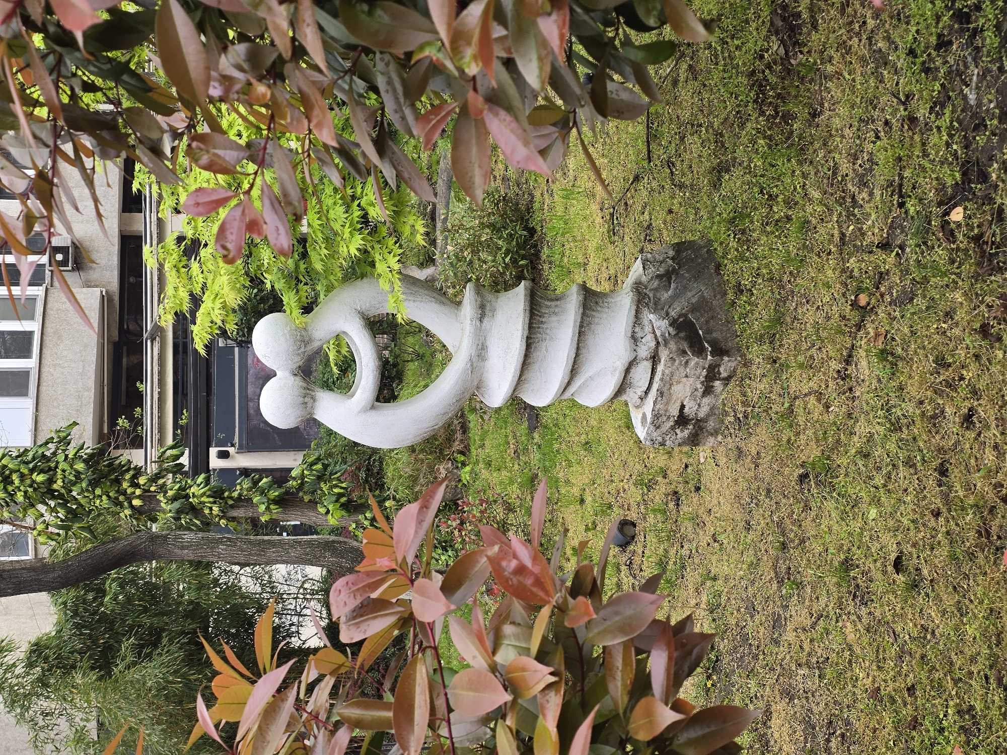 A white sculpture of two intertwined figures stands on a grassy lawn in an urban park. The sculpture is made of concrete and has a smooth, white finish. The lawn is green and slightly overgrown. The sculpture is surrounded by trees and bushes. In the background, a building can be seen. The location of the sculpture is in the city of Unknown, Unknown.