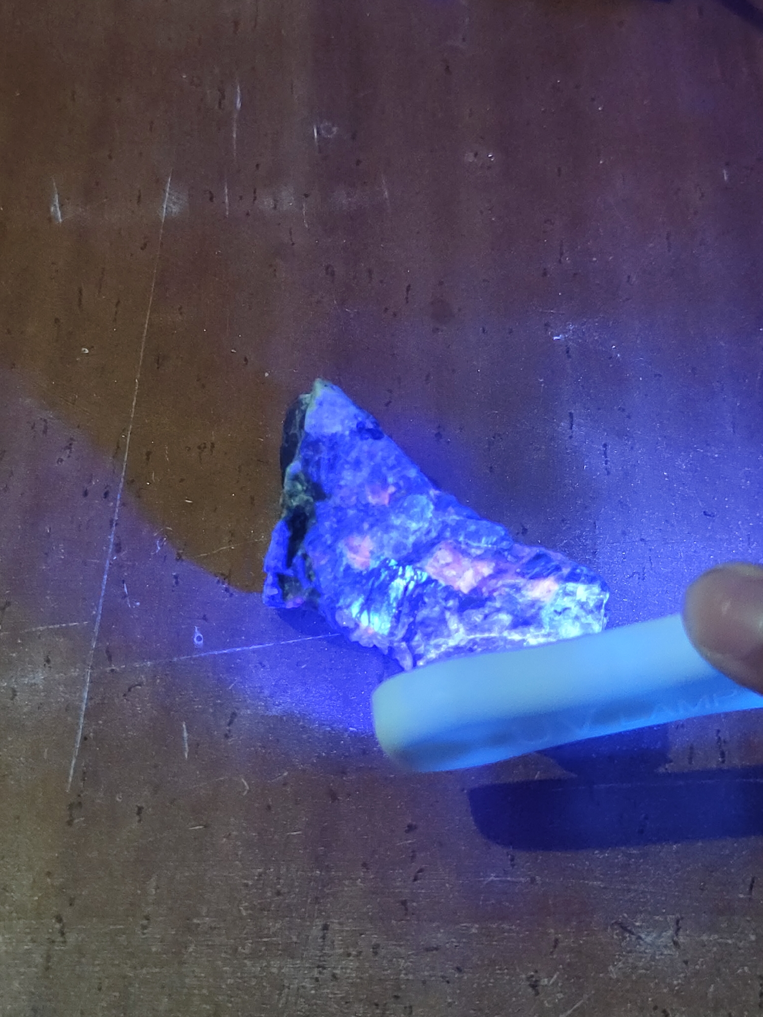 Within a dimly lit setting in Davao City, Philippines, the image captures a mineral specimen exhibiting strong fluorescence under ultraviolet (UV) light. The irregular, rough-textured specimen, positioned centrally, glows intensely with vibrant blue and purple hues, interspersed with distinct patches of orange and pink fluorescence. A white, elongated object, consistent with a UV flashlight, is held by a human hand, with the tips of a thumb and forefinger visible in the lower right corner, actively directing the light onto the specimen. The immediate surface beneath the specimen and flashlight is a dark brown, possibly wooden, material characterized by numerous visible scratches, scuffs, and small indentations, indicative of wear. The UV light casts a distinct dark shadow from the flashlight onto this worn surface.