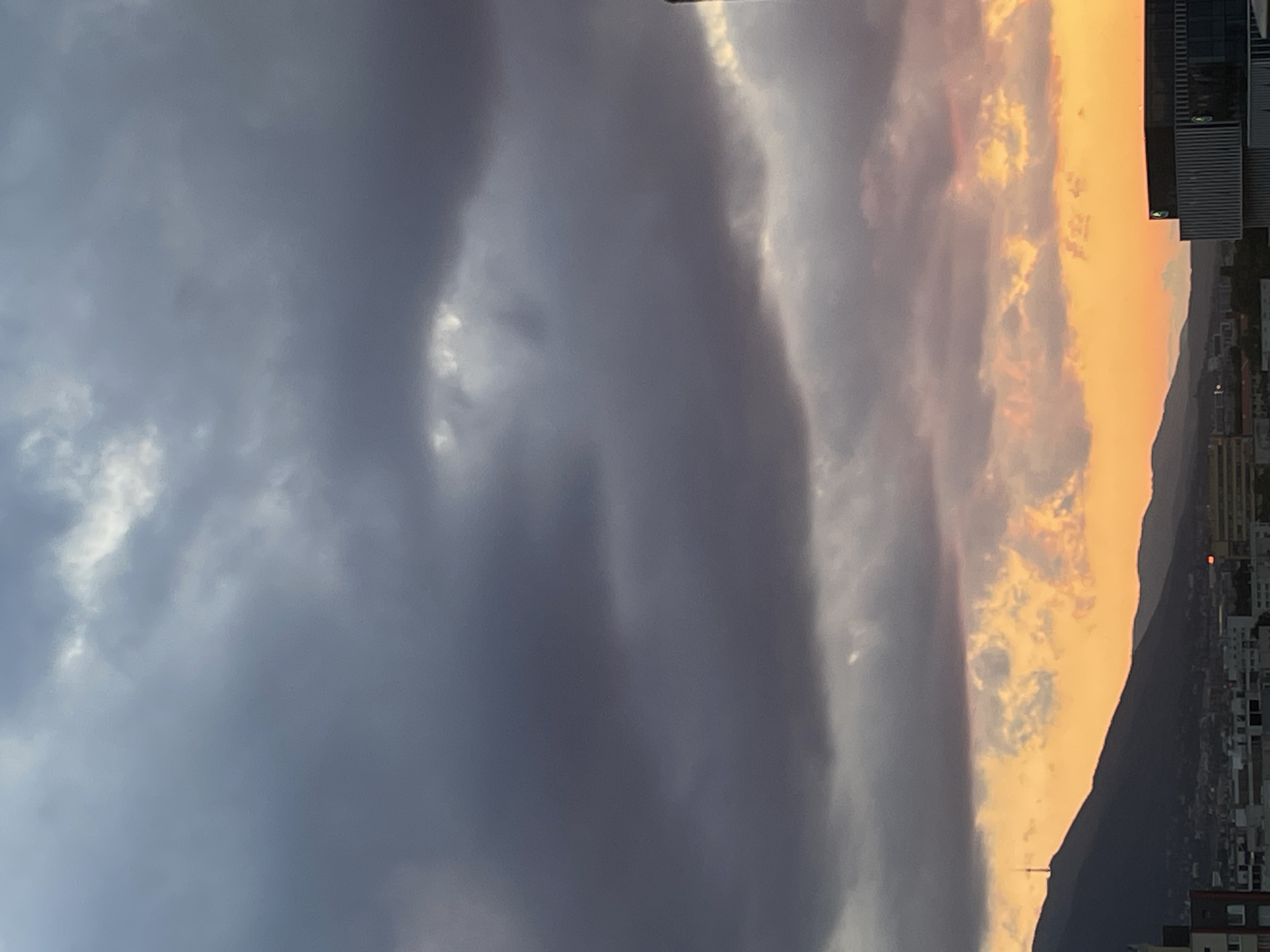 This image captures a sunset over the city of Sofia, Bulgaria, with Vitosha Mountain dominating the background. The upper two-thirds of the frame are filled with dense, dark grey to bluish-grey clouds, exhibiting some internal lighter patches. The lower third of the sky presents a dramatic sunset, characterized by vibrant orange, yellow, and reddish hues near the horizon, indicating the sun is low or just below it.
In the midground, the dark silhouette of Vitosha Mountain stretches across the left and central portions, its peak featuring the distinct Vitosha TV Tower, a slender communications structure. Below the mountain, the urban landscape of Sofia unfolds, showing a densely packed collection of buildings. These structures, mostly in shadowed tones, vary in height and architectural style, with a modern building featuring a dark, rectangular facade and a lighter, ridged section prominent in the lower right foreground. No specific people or discernible actions are visible within the frame. The overall lighting is dim, characteristic of dusk, with strong contrast between the dark foreground elements and the brightly illuminated horizon.