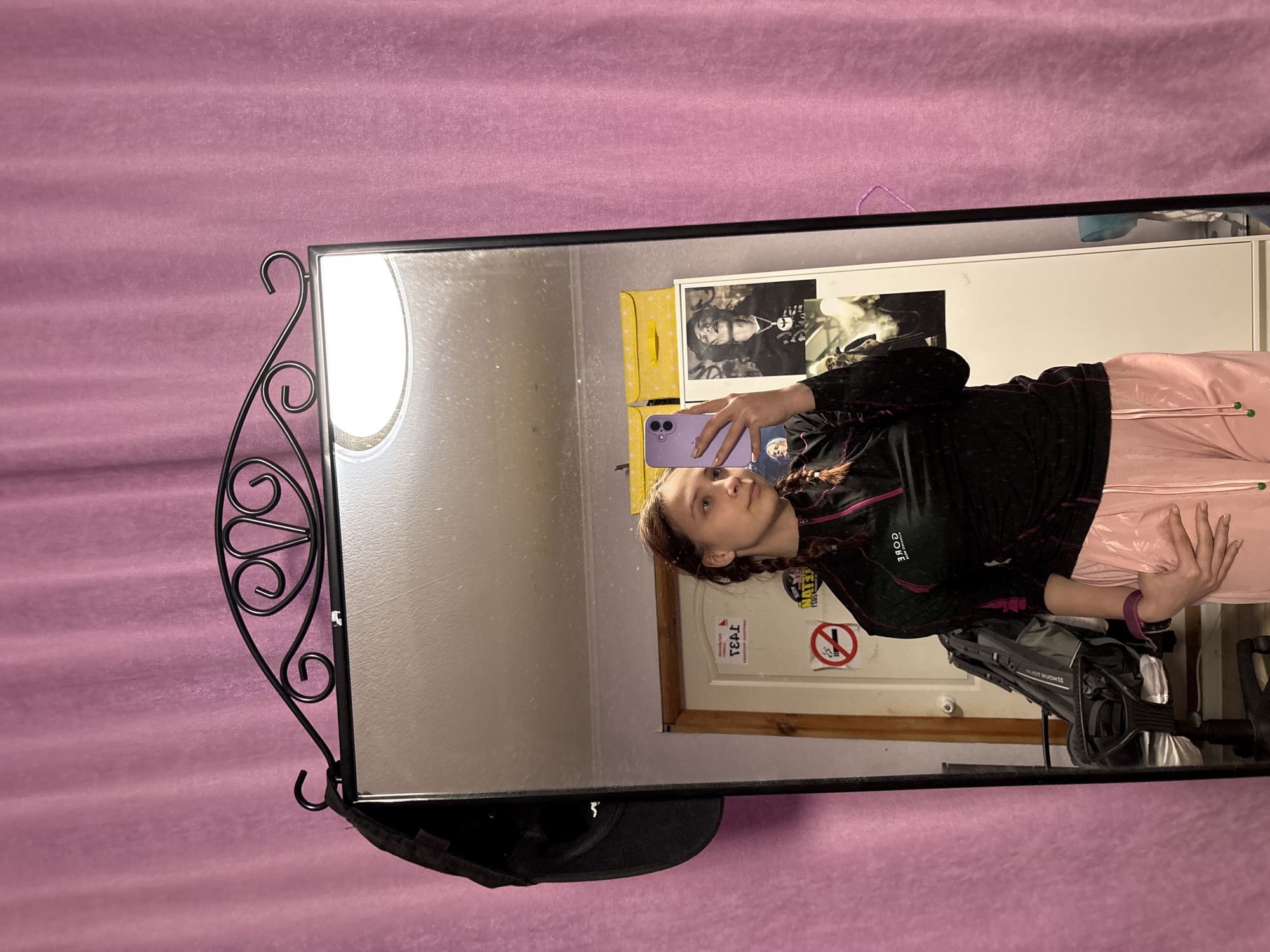 A selfie of a person with long braided hair, wearing a black athletic jacket and light pink pants. They are holding a purple phone in their left hand, taking a picture in a mirror with a decorative black metal frame. The mirror reflects a room with a white door, a "no smoking" sign, and a poster of a person. A black baseball cap hangs on the lower edge of the mirror. A pink curtain is visible in the background. There is no information in the image to verify it is taken in the city of Прикамский, Russia.