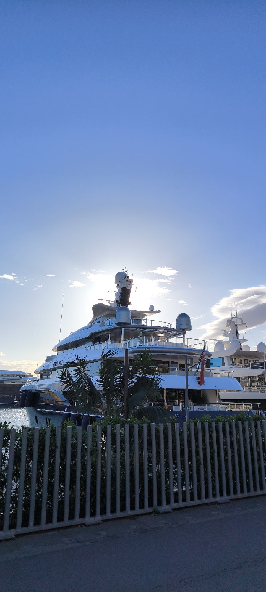 The image shows a large, multi-tiered yacht docked in a marina. The yacht is predominantly white and silver, with reflective surfaces and satellite domes on its upper decks. A small red flag is visible. The sun is shining brightly behind the yacht, creating a glare and silhouetting parts of the vessel. In the foreground, there is a metal fence and some greenery, partially obscuring the view. The marina setting includes other yachts visible in the background. Considering the architectural style of the surrounding yachts and the overall urban feel, it’s plausible that the photograph was taken in Barcelona, Spain, specifically in Port Vell, a harbor located at the edge of Barcelona's old city. There are no people visible in the image.