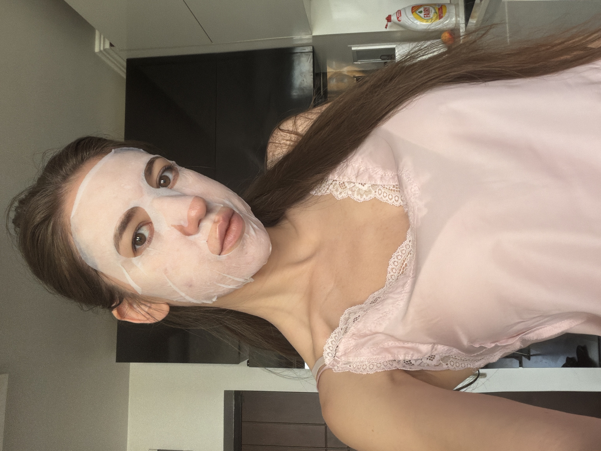 A young woman is in a kitchen applying a facial mask. The woman has long brown hair, which is pulled back from her face, and she is wearing a light pink camisole with lace trim. The kitchen appears to be modern, with dark cabinetry and white countertops. A bottle of dish soap is visible on the counter. The image is a selfie, taken from a slightly low angle.