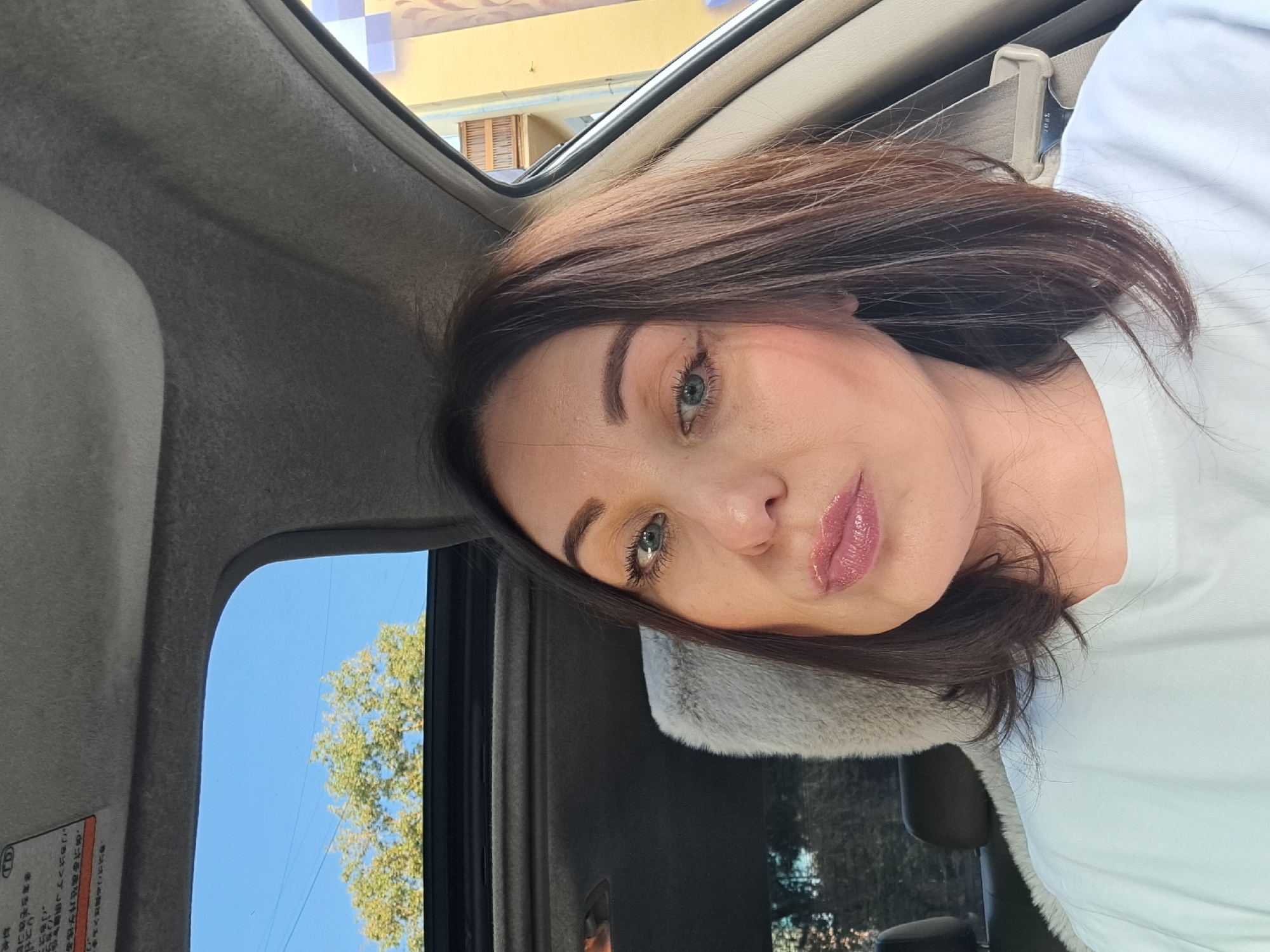 A woman with brown hair and blue eyes is taking a selfie in the driver's seat of a car. She is wearing a white t-shirt. The car has a sunroof, and through it, a tree and blue sky are visible. In the background, a building with yellow and blue checkered elements can be seen. The image is taken outdoors on a sunny day.