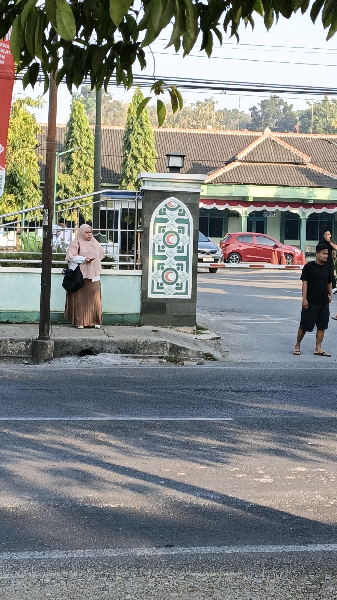 In Kuwasen, Indonesia, on a street with a sidewalk and asphalt road, a woman wearing a hijab and a long skirt stands on the sidewalk, holding a phone. Beside her is a black bag. She is standing in front of a pillar with Islamic-style ornamentation. To her left, a metal fence and some trees are visible. Behind her, a building with a red and green facade and a tiled roof can be seen. In the street, a red car is parked, and a man in a black t-shirt and shorts is walking away from the camera, wearing sandals. The scene appears to be set in the late afternoon, with shadows cast across the road by unseen trees.