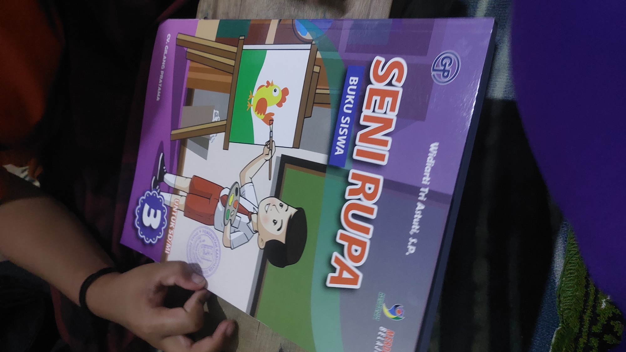 The image shows a close-up of a children's book titled "SENI RUPA" (Art) for grade 3 students in Indonesia. A person's hand, with a black wristband, is visible in the upper left corner, holding the book. The book cover features an illustration of a young boy painting a yellow bird on an easel. The book is oriented upside down from the viewer's perspective. Text on the book includes "UNTUK SD/MI," "BUKU SISWA," "CV. CILANG PRATAMA," and "Widjarti Tri Astuti, S.P." There are also logos of "MEBOS SEJAHTERA" and "GP." The book is resting on a wooden surface. The background is dark and slightly out of focus, suggesting an indoor setting. There is no visible information within the image to determine the specific location within Jatirogo, Indonesia.
