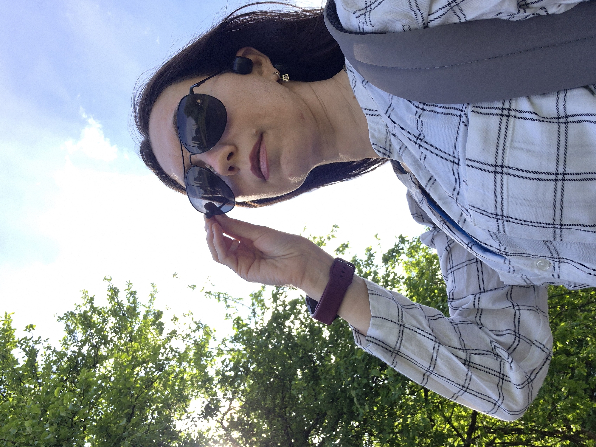 The image is a low-angle shot of a woman wearing sunglasses, a plaid shirt, and a burgundy smartwatch. She is holding the rim of her sunglasses in her right hand, partially obscuring her face. She has dark hair and is wearing wireless earbuds. A gray strap crosses her body diagonally. The background consists of green trees illuminated by sunlight and a blue sky. The location is Unknown, Unknown, but could possibly be a park or outdoor area.