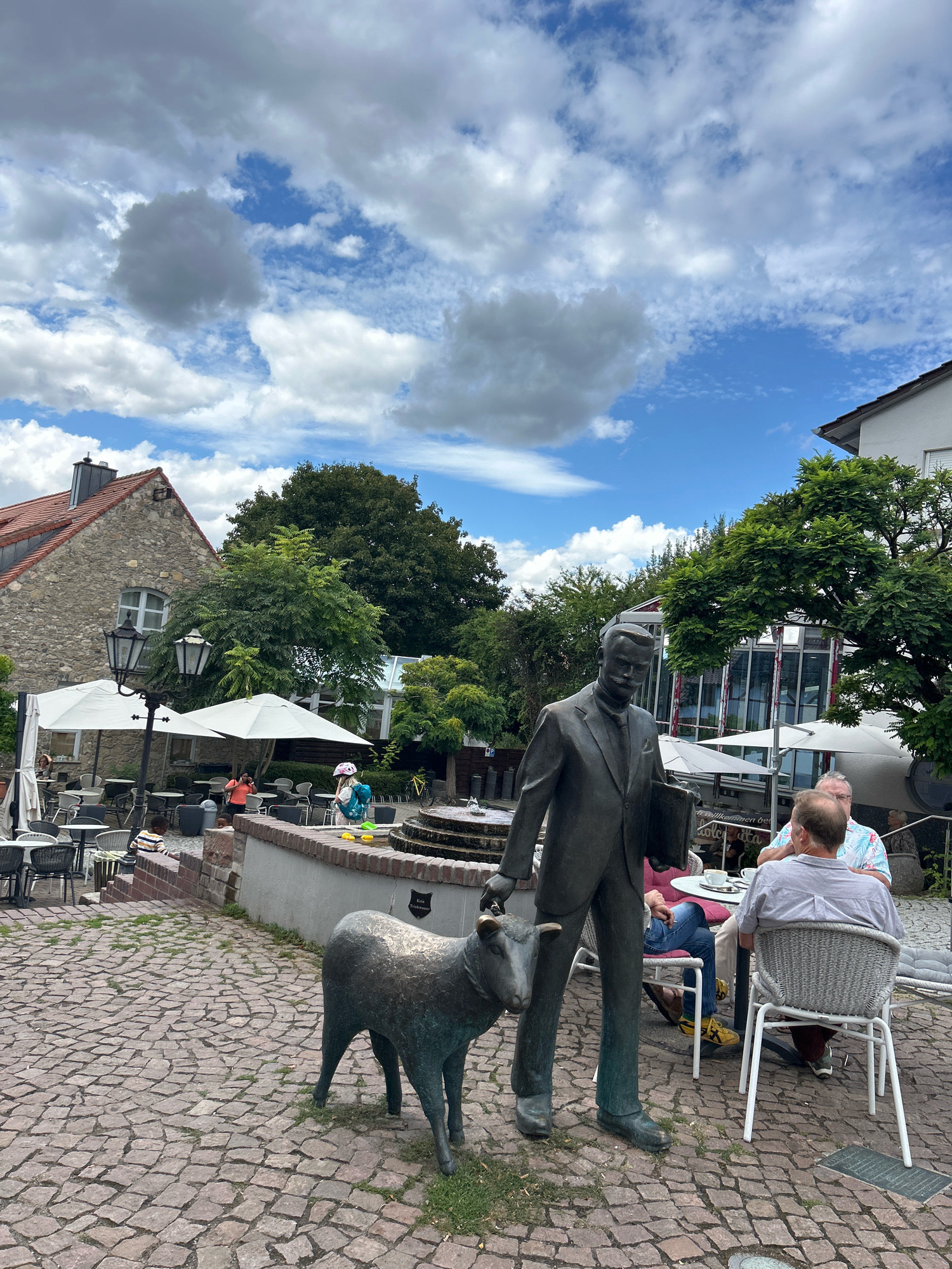 In the city of Unknown, Unknown, on a cloudy day with patches of blue sky, a bronze statue of a man in a suit, holding a book, stands on a cobblestone plaza next to a bronze statue of a sheep. The man is looking down at the sheep, and the sheep is looking forward. Behind the statues, an outdoor cafe is set up with white umbrellas and tables. Several people are seated at the tables, some conversing and others looking at their phones. A stone building with a red tiled roof and a large tree are visible in the background, as are other cafe tables and chairs. The scene appears to be a peaceful outdoor setting in a European city.